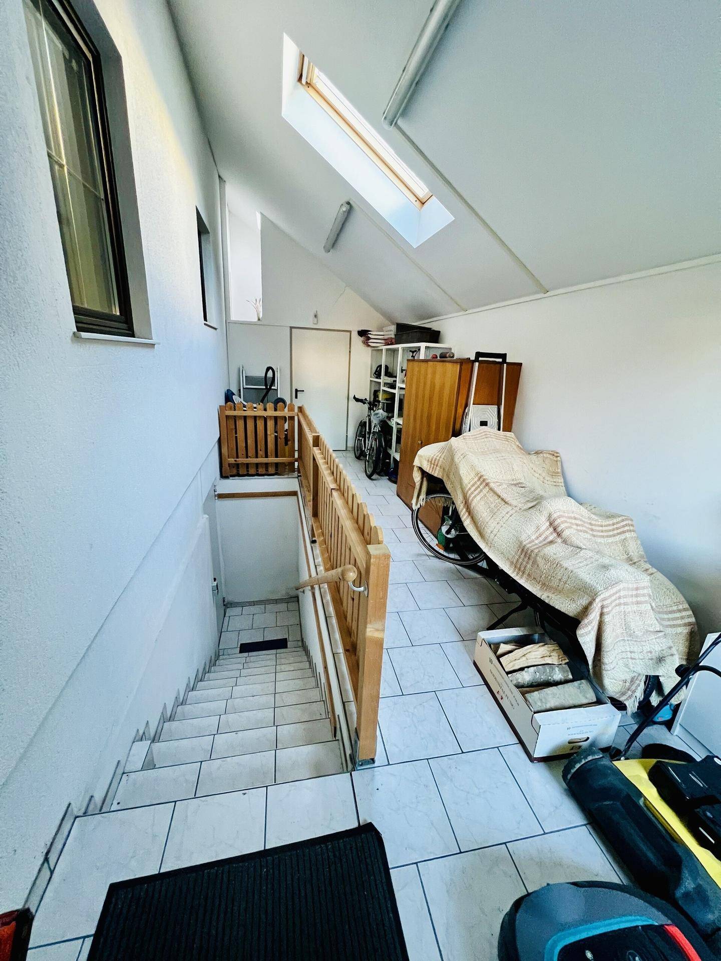 Treppenhaus mit Fliesenboden, Holzgeländer und Oberlicht, das Tageslicht hereinlässt.