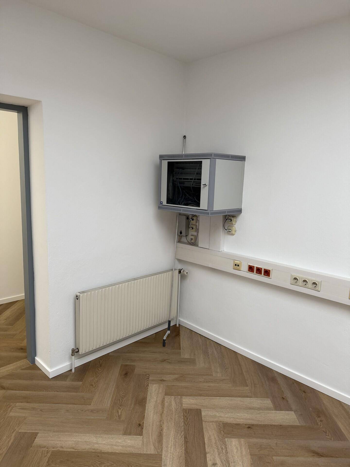 Ecke eines Büroraums mit Fischgrätparkett, Heizkörper und einem an der Wand montierten Schrank.