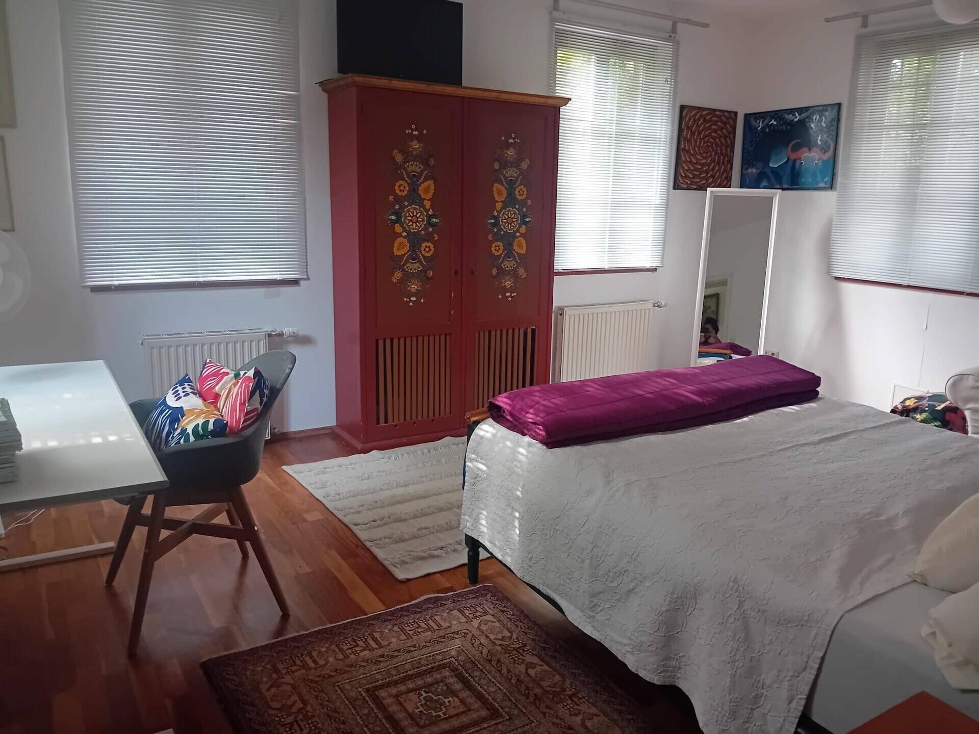 Schlafzimmer mit Holzboden, Schreibtisch und farbenfrohem Schrank, helle Fenster.