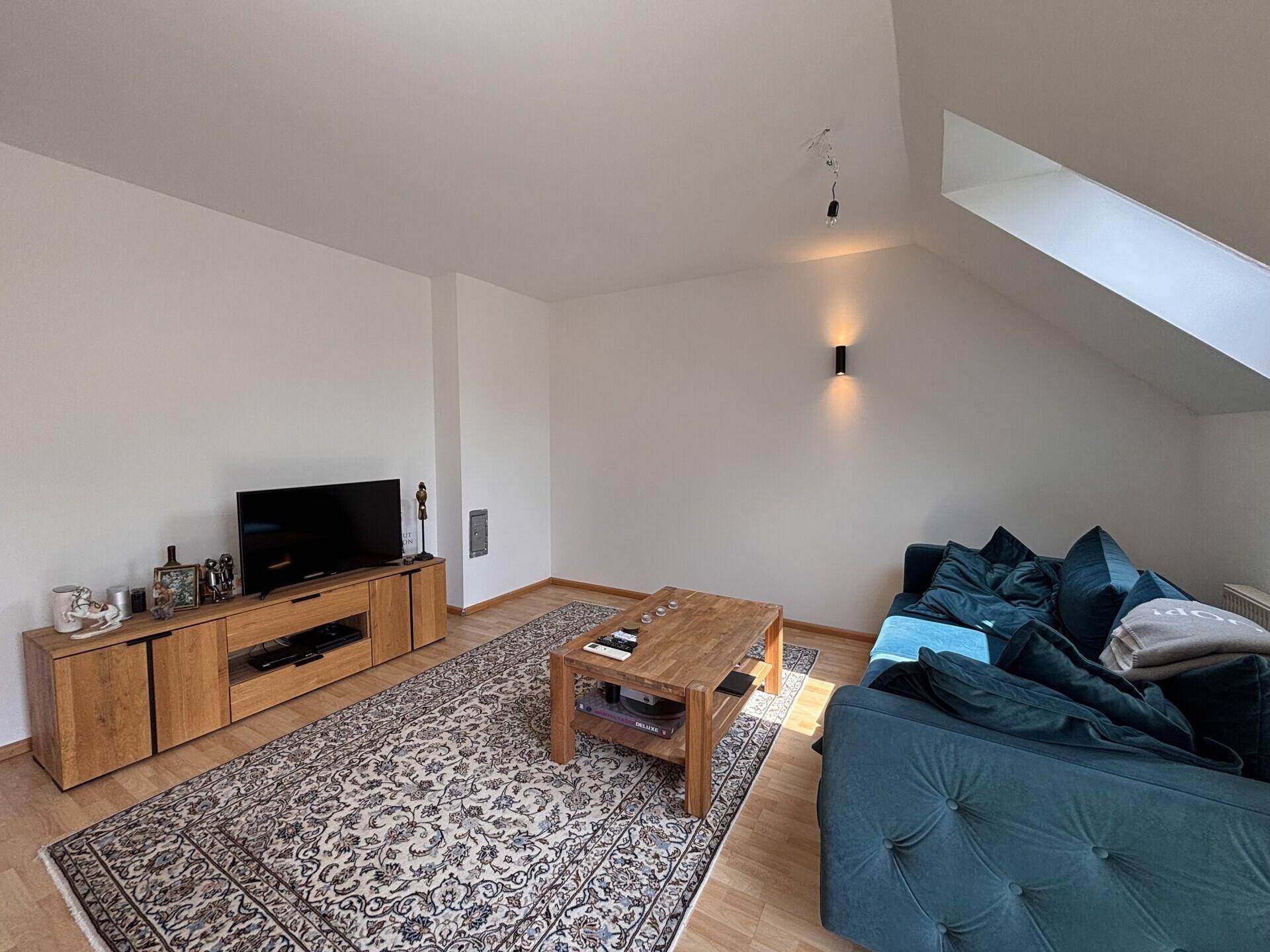 Gemütliches Wohnzimmer mit dunkelgrünem Sofa, Holztisch und Teppich unter schräger Decke.