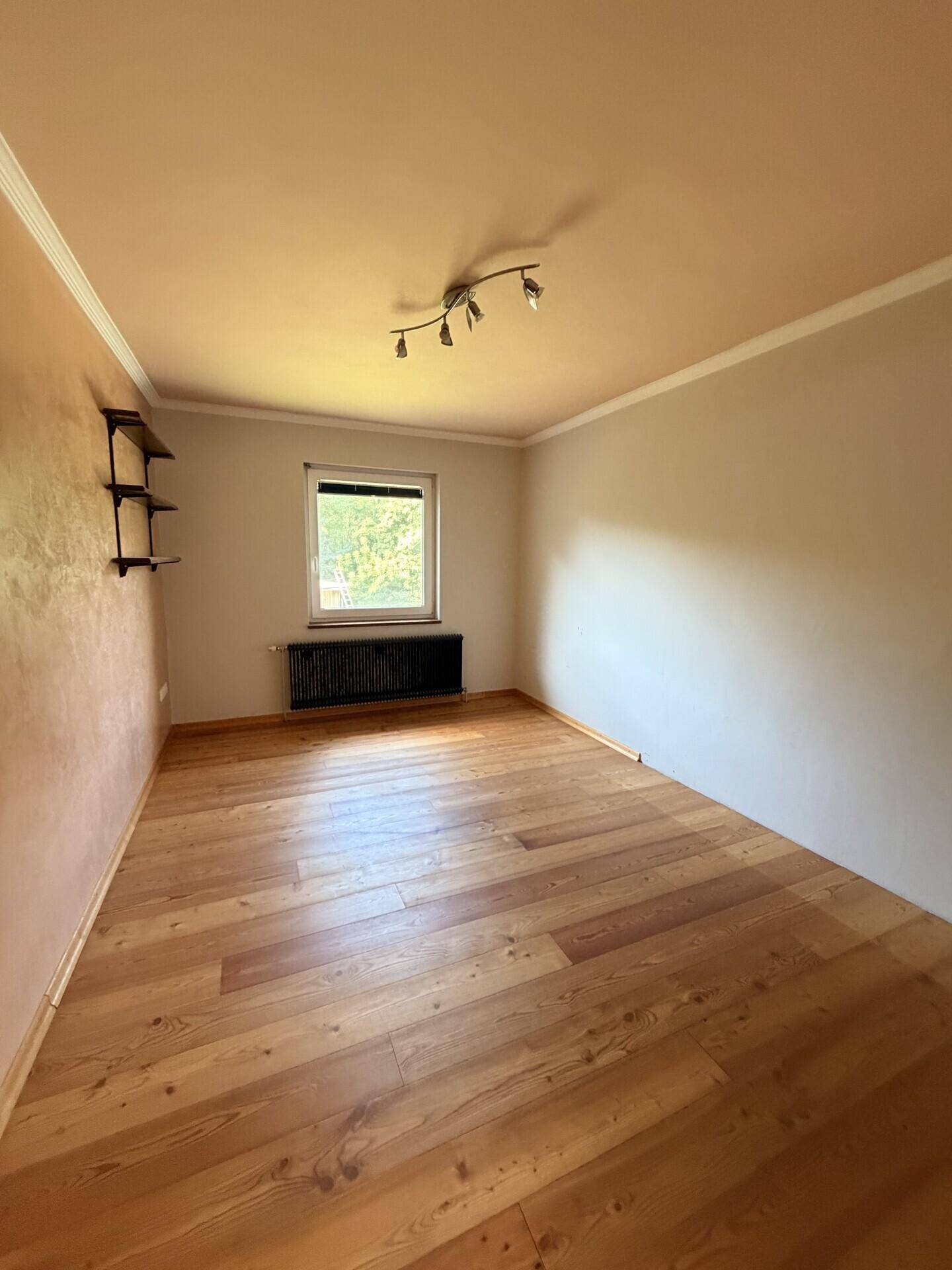 Helles Schlafzimmer mit Holzboden, beigen Wänden und einem Fenster mit Blick ins Grüne.