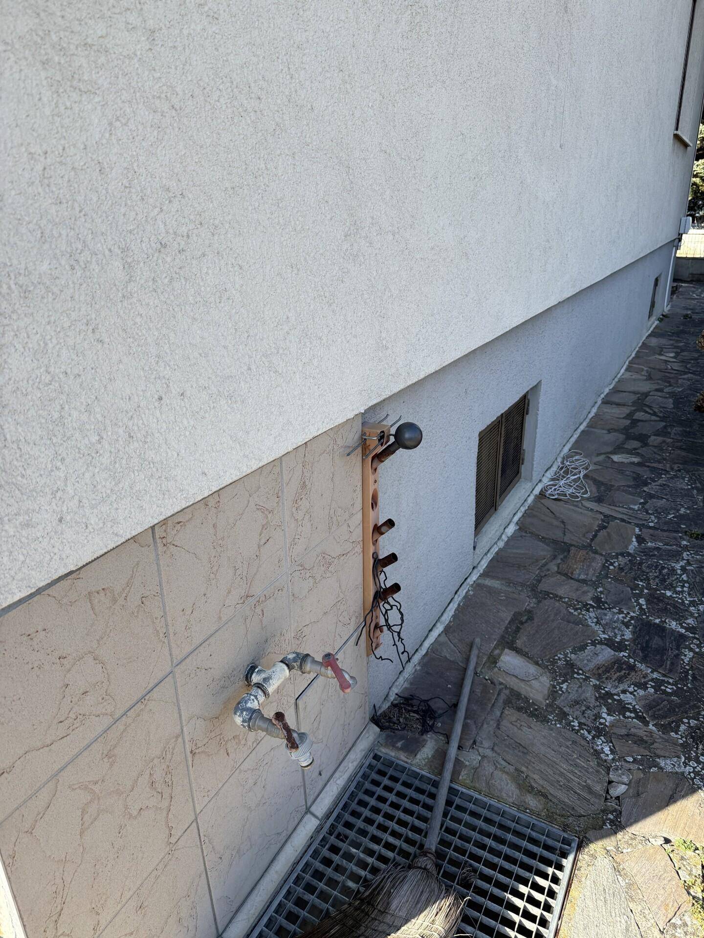 Hauswand mit sichtbaren Wasseranschlüssen und einem Entwässerungsrost am Boden.