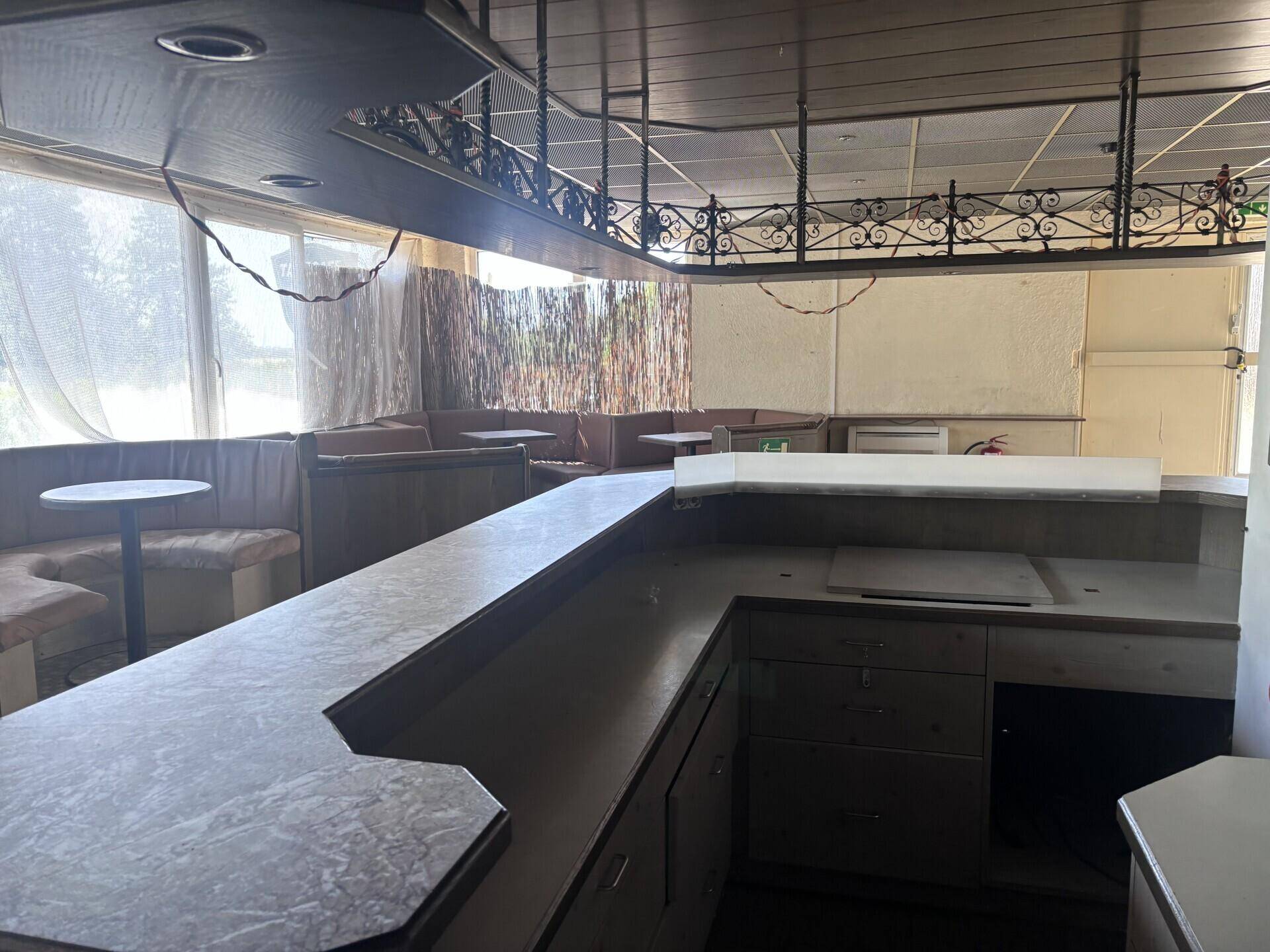Innenansicht einer Bar mit langer Theke, gepolsterten Sitzbänken und Fenstern mit Vorhängen.