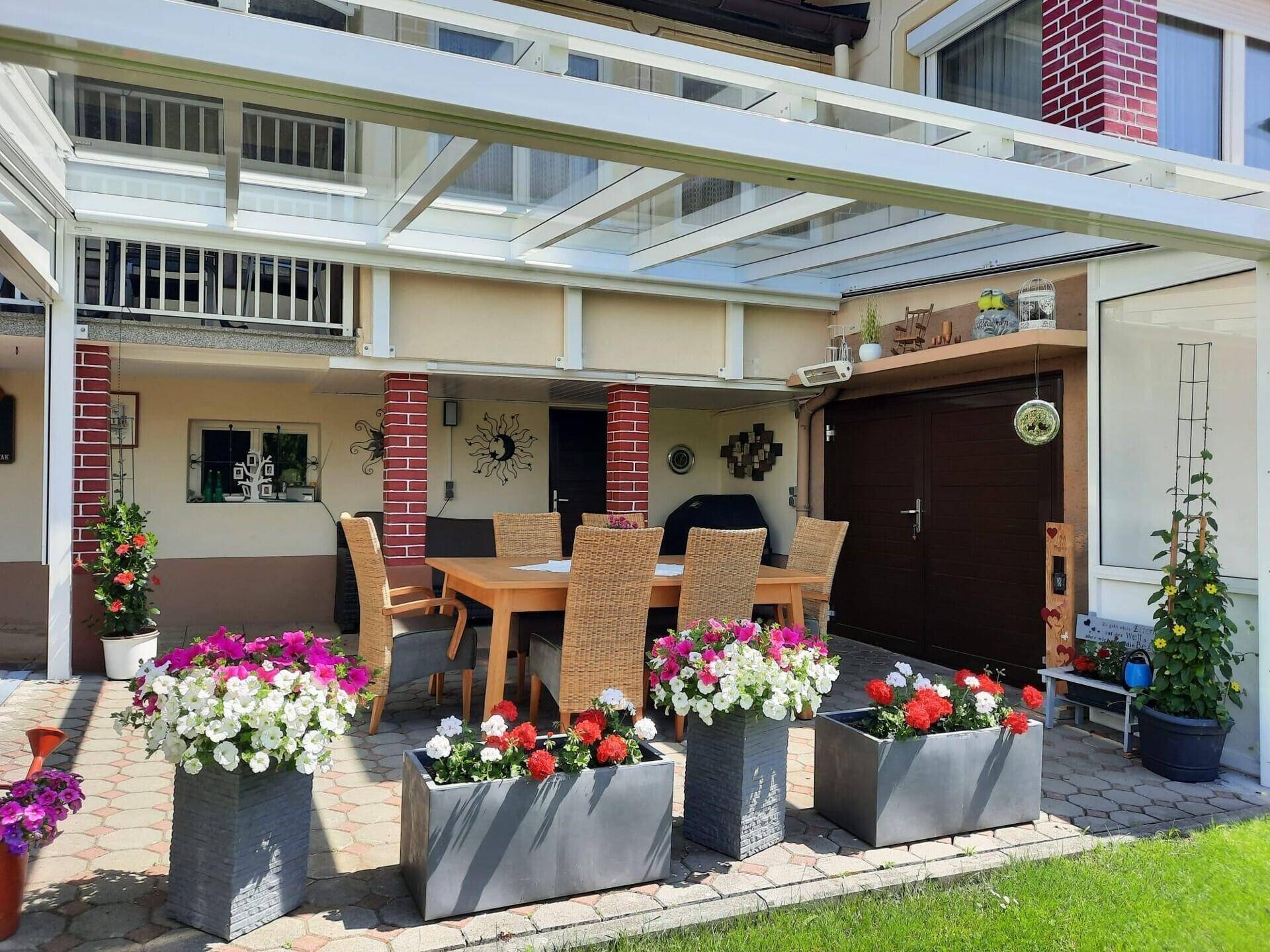 Überdachte Terrasse mit Esstisch und Blumenkübeln, angrenzend an die Hausfassade und Garage.