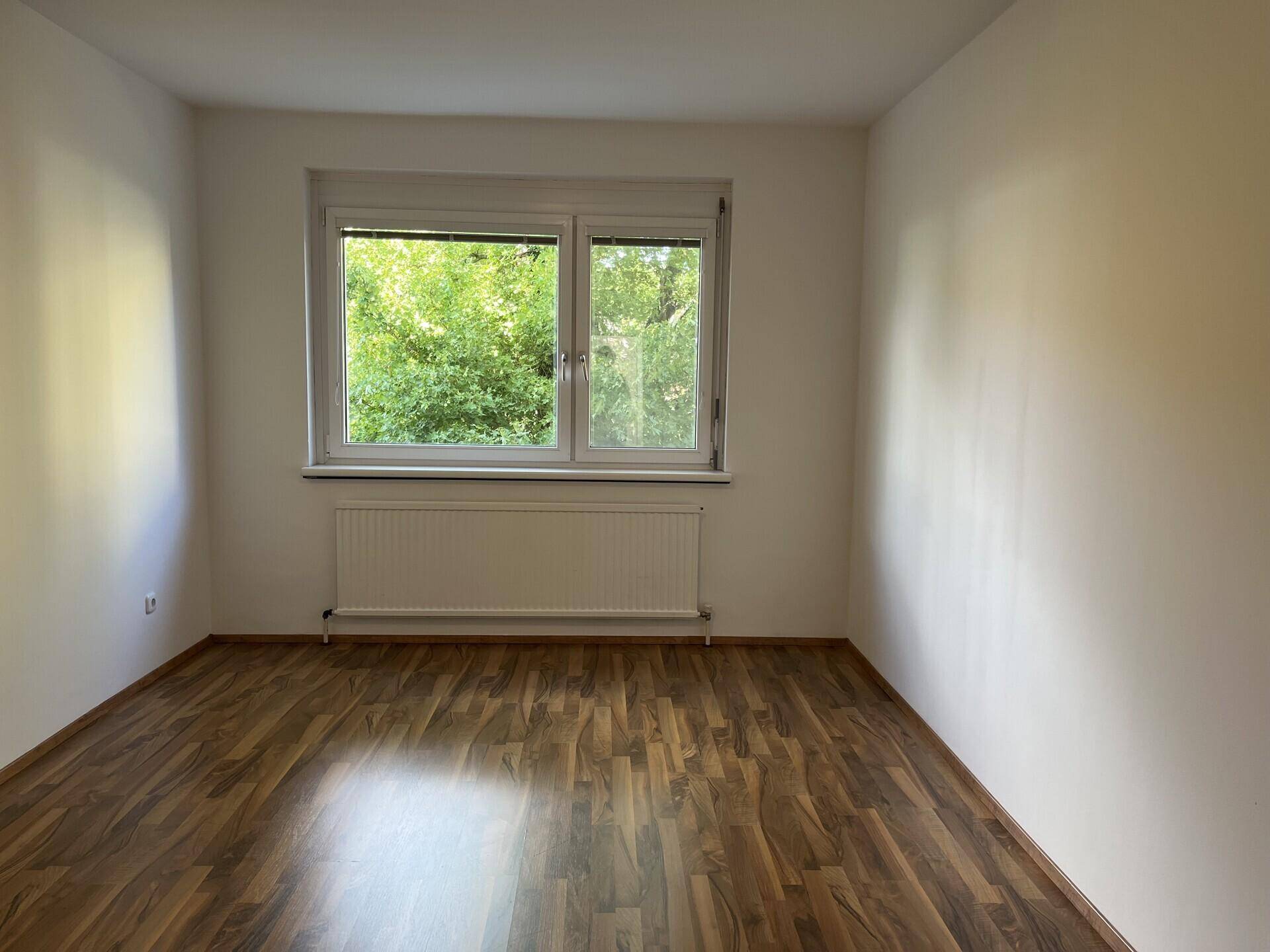 Heller Raum mit dunklem Laminatboden und großem Fenster, das einen schönen Grünblick bietet.
