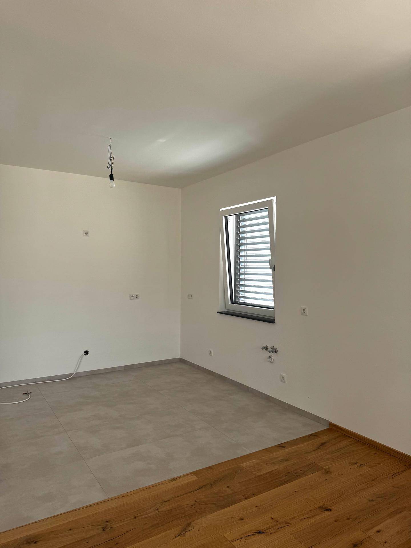 Heller Raum mit weiß gestrichenen Wänden, einem Fenster mit Jalousien und gemischtem Bodenbelag.