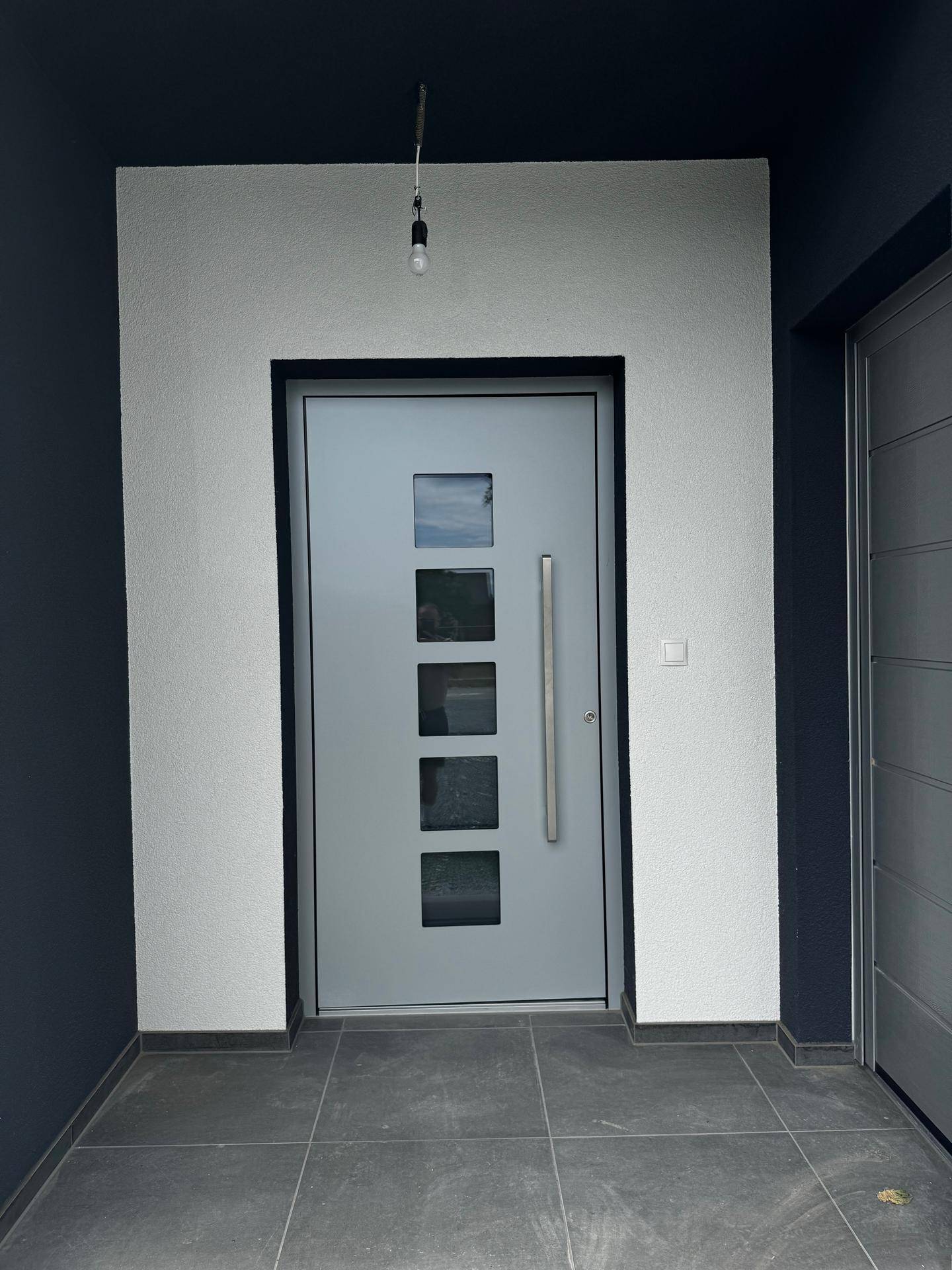 Moderner Hauseingang mit einer grauen Tür, Glaseinsätzen und dunklen Fliesen im Eingangsbereich.