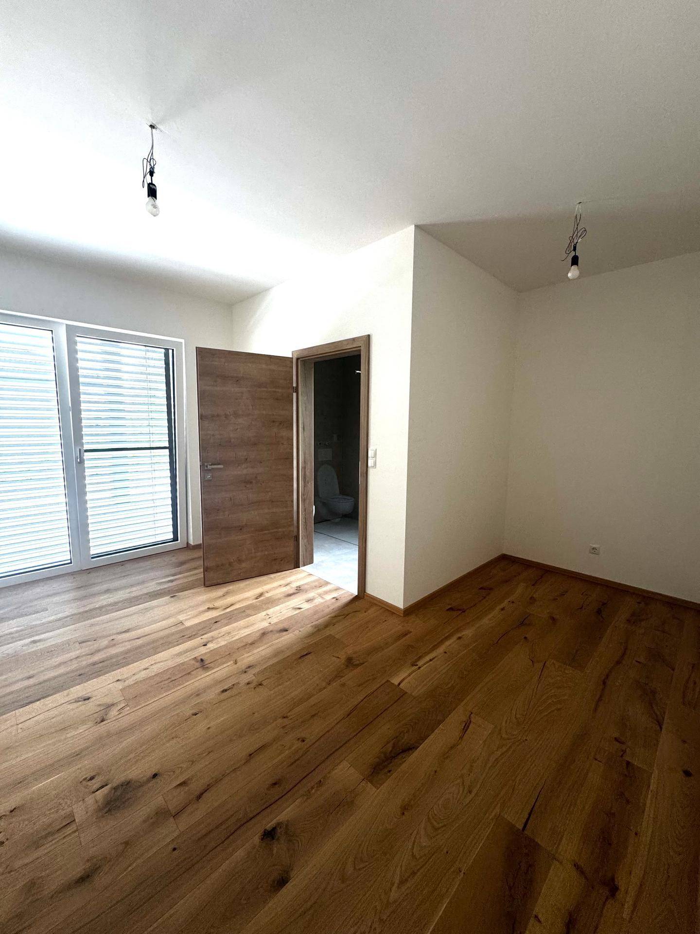 Helles Schlafzimmer mit Holzboden und großen Fenstern, die viel Tageslicht hereinlassen.