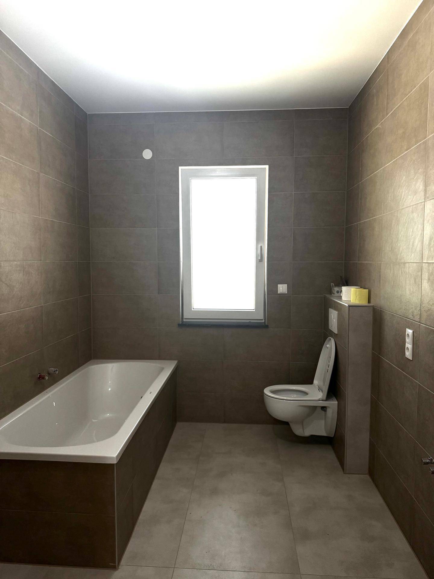 Modernes Badezimmer mit braun-grauen Fliesen, einer Badewanne, wandhängendem WC und Fenster.