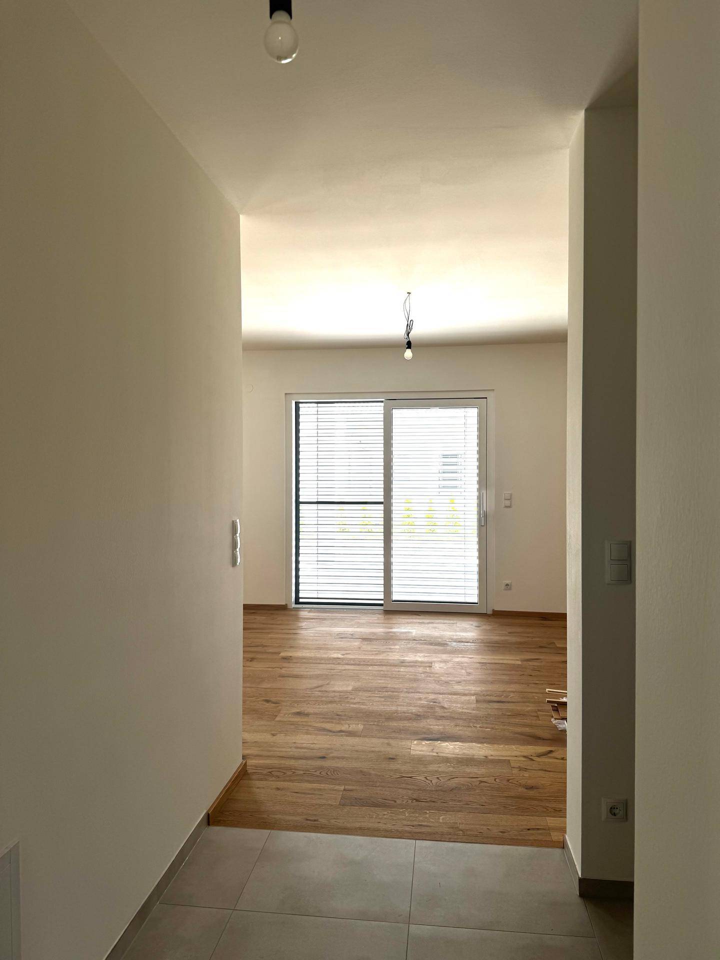 Langer Flur mit grauen Fliesen, der zu einem hellen Wohnbereich mit Holzboden und großen Fenstern führt.