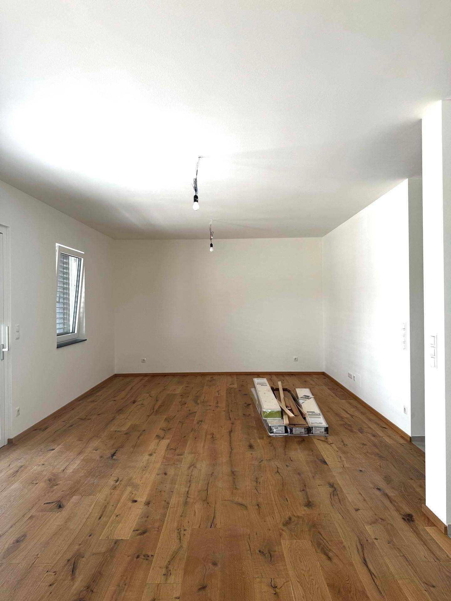 Geräumiges, helles Wohnzimmer mit neuem Holzboden, weißen Wänden und einem Fenster mit Jalousien.