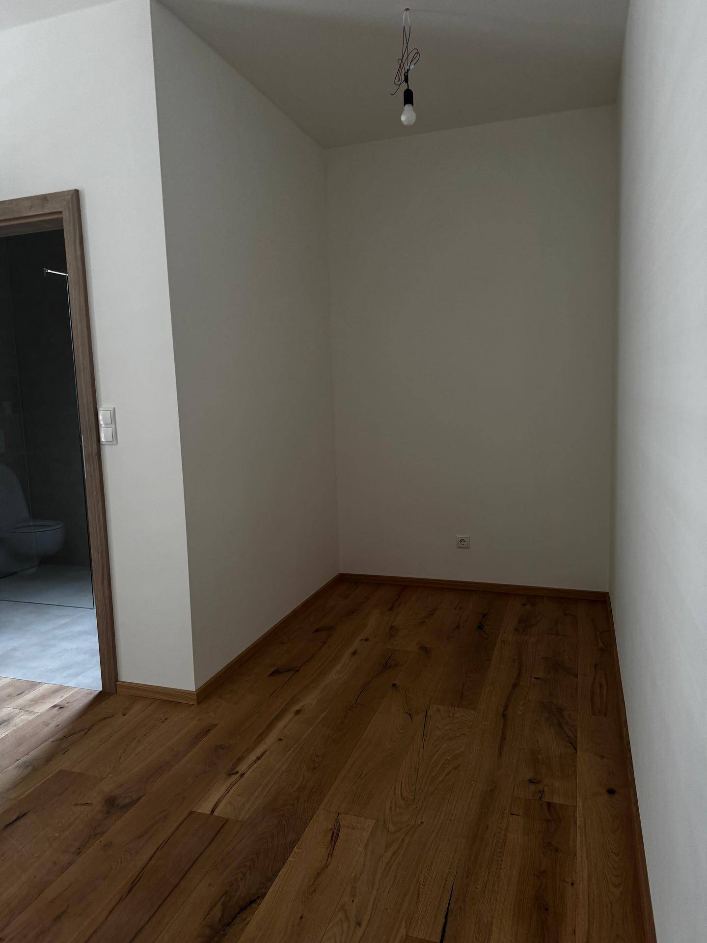 Heller Raum mit Holzboden und weiß gestrichenen Wänden, angrenzend an ein Badezimmer.