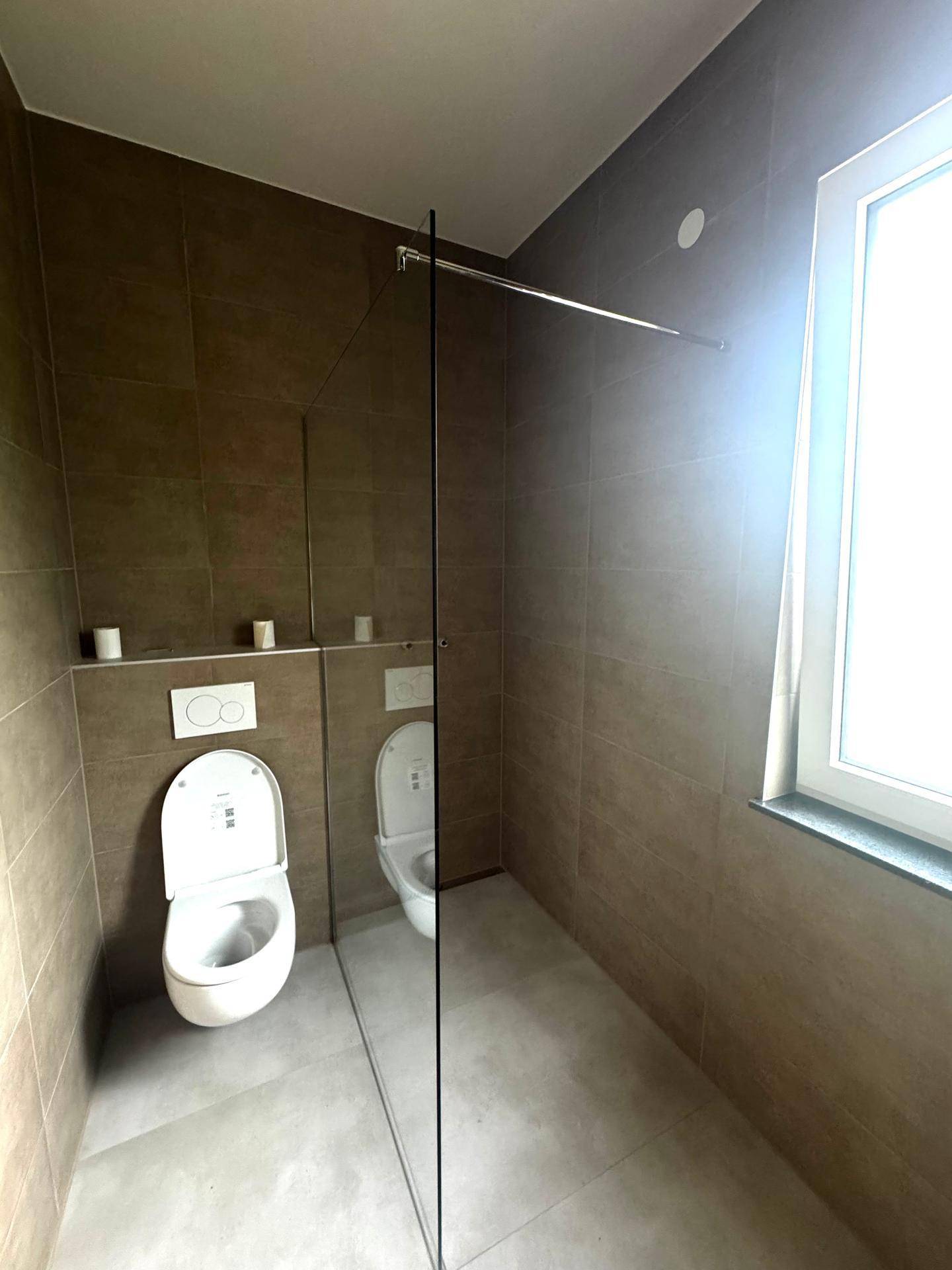 Modernes Badezimmer mit Duschbereich, wandhängendem WC, braun-grauen Fliesen und Fenster.