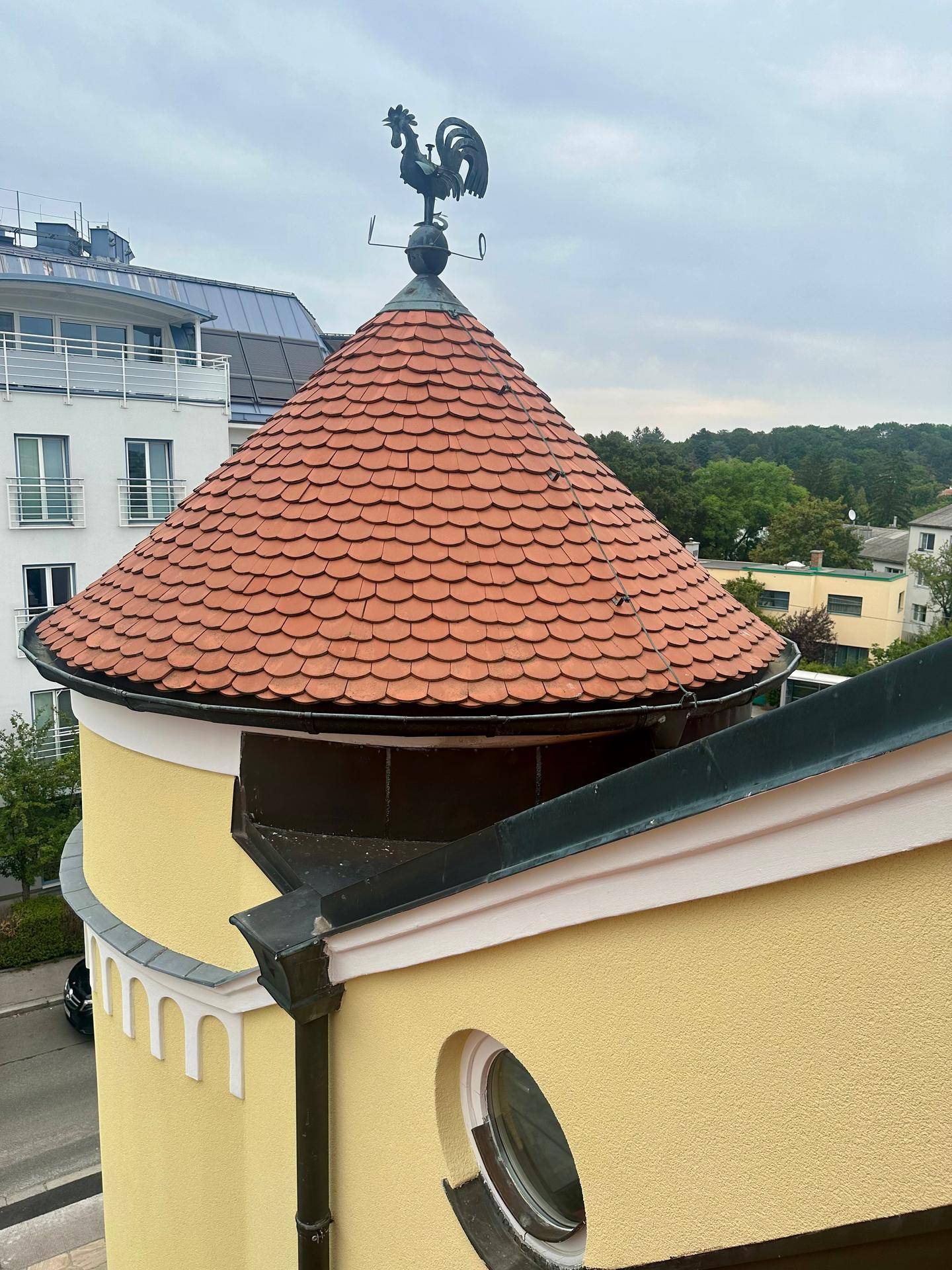Nahaufnahme des Turmdachs mit Wetterhahn und Ziegeln, umgeben von Bäumen und anderen Gebäuden.