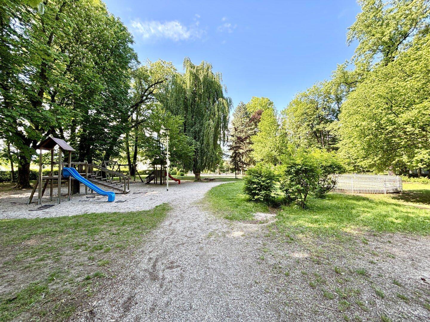 Ein Spielplatz mit Rutsche und Schaukeln, umgeben von hohen Bäumen in einem Park.