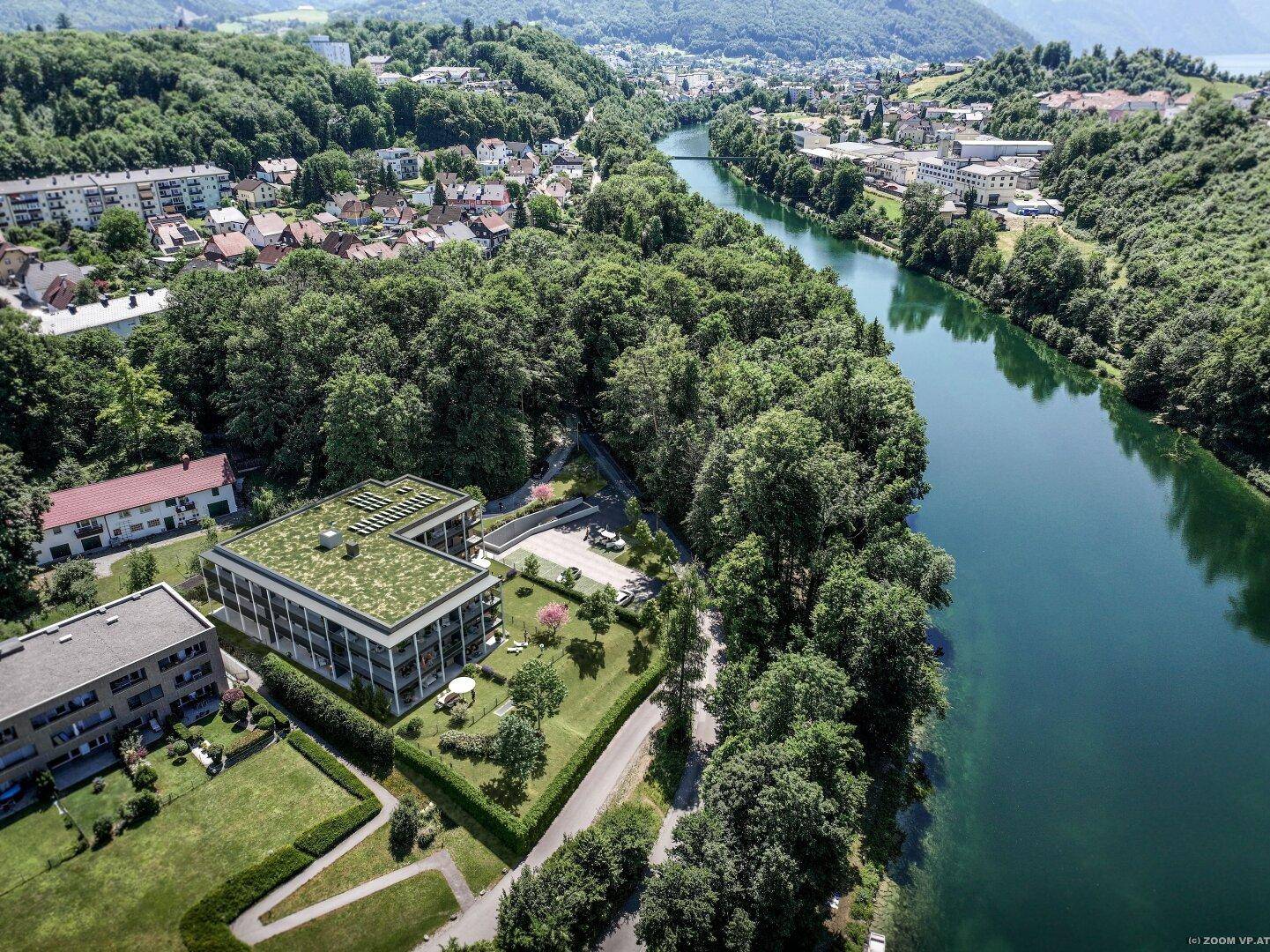 Luftaufnahme einer modernen Wohnanlage mit Gründach direkt am Fluss, umgeben von viel Grün.