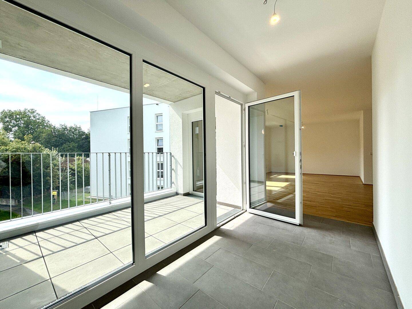 Heller Innenraum mit großflächigen Fenstern und Zugang zu einem modernen Balkon mit grüner Aussicht.