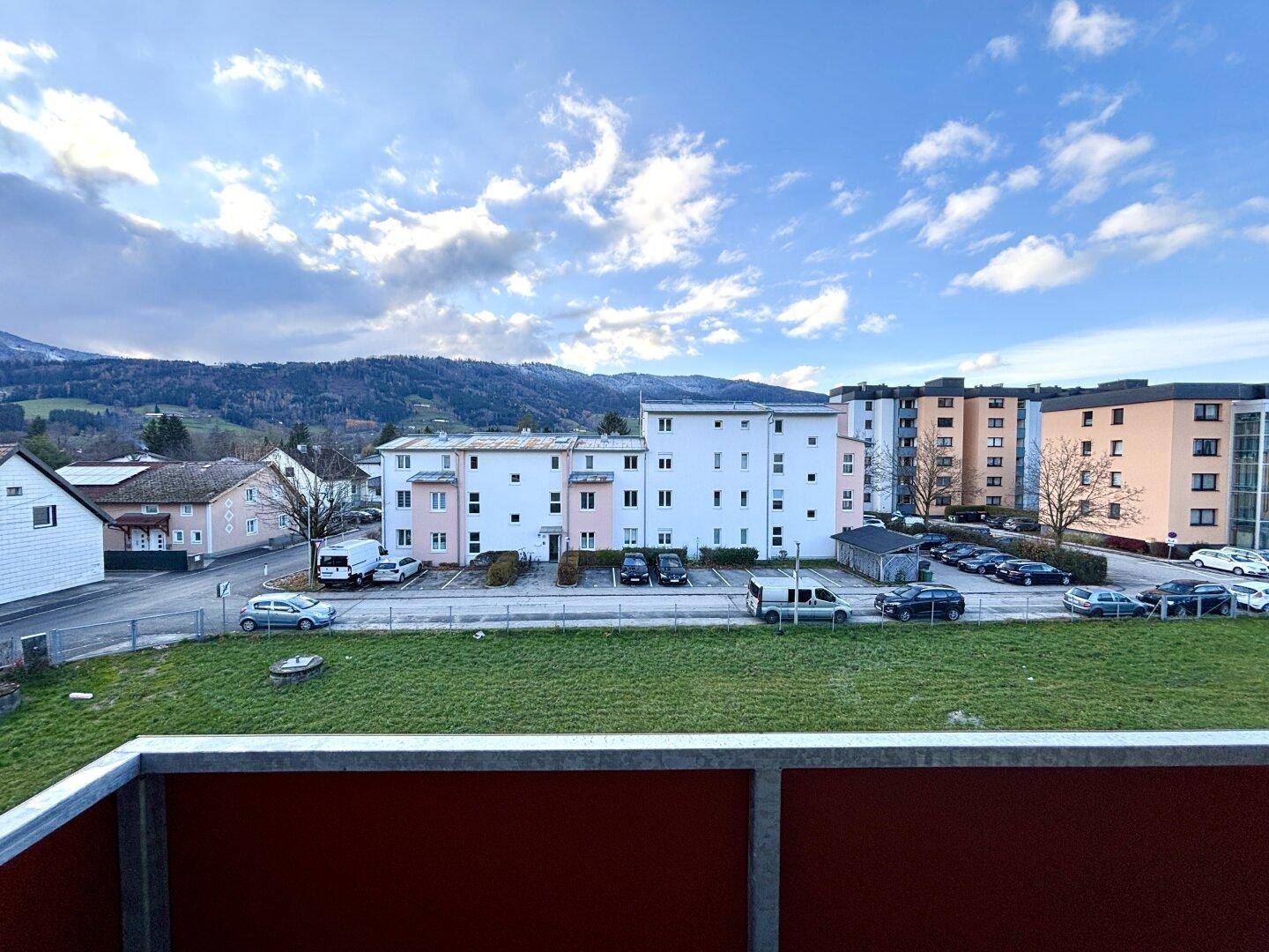 Blick von einem Balkon auf eine Wohnsiedlung mit grünen Flächen und Bergen im Hintergrund unter bewölktem Himmel.