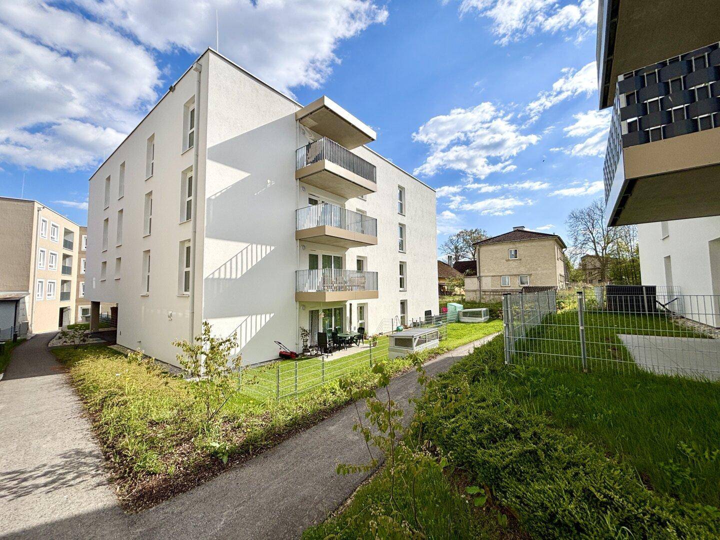 Modernes Mehrfamilienhaus mit weißer Fassade, Balkonen und einer möblierten Terrasse im Erdgeschoss.