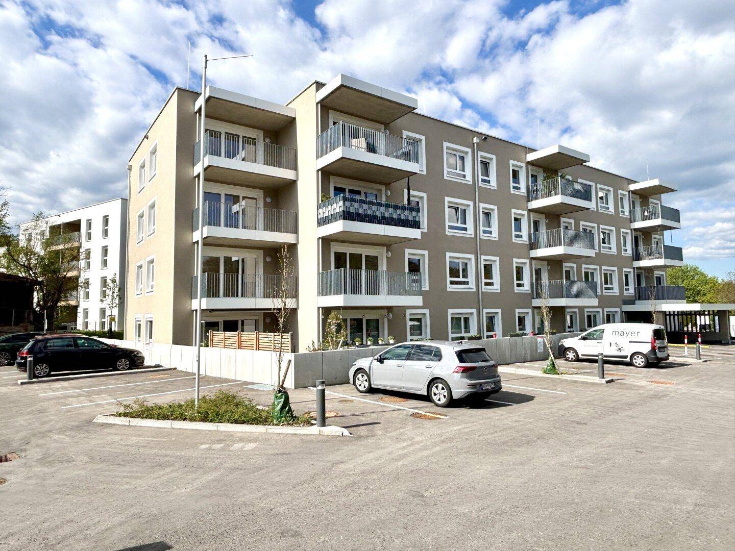 Modernes Mehrfamilienhaus mit mehreren Balkonen und Außenparkplätzen unter bewölktem Himmel.