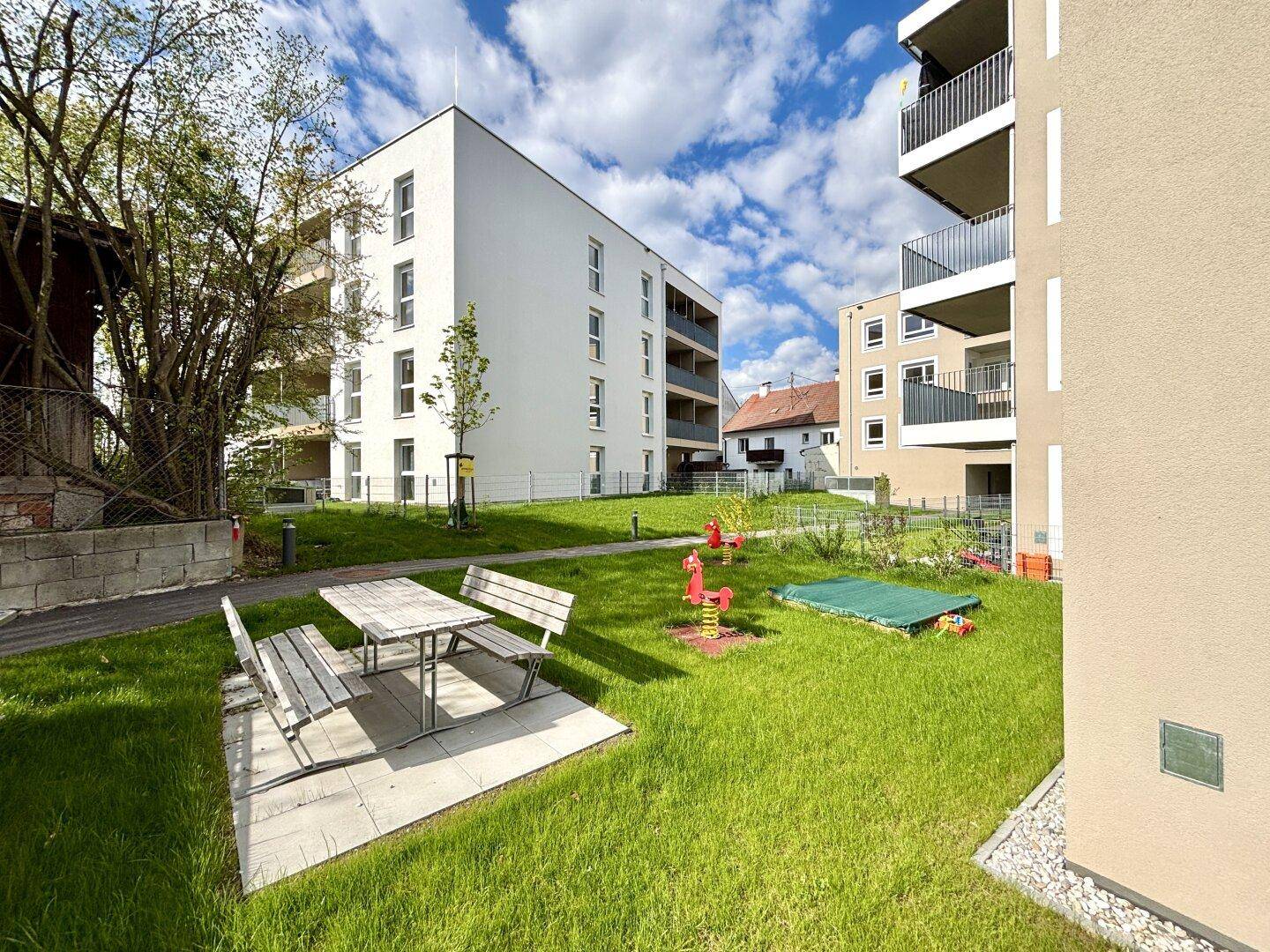 Grüner Gemeinschaftsgarten mit Picknicktisch, Spielplatz und modernen Wohngebäuden im Hintergrund.