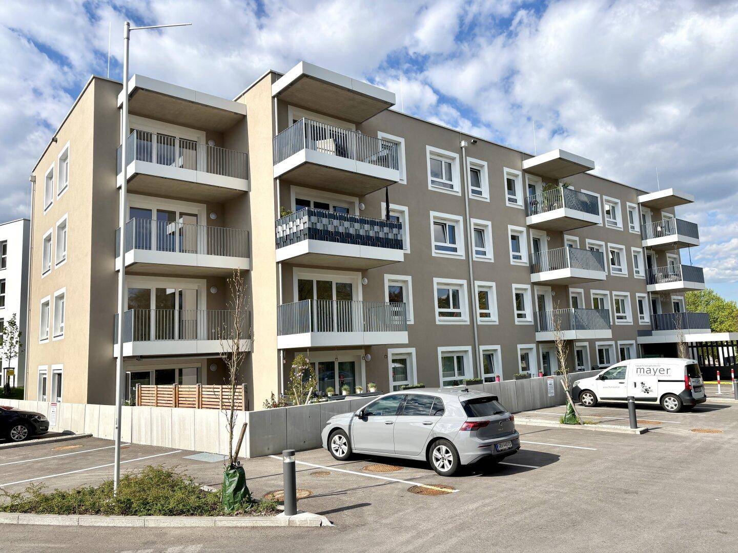 Modernes Mehrfamilienhaus mit mehreren Balkonen und Außenparkplätzen unter bewölktem Himmel.