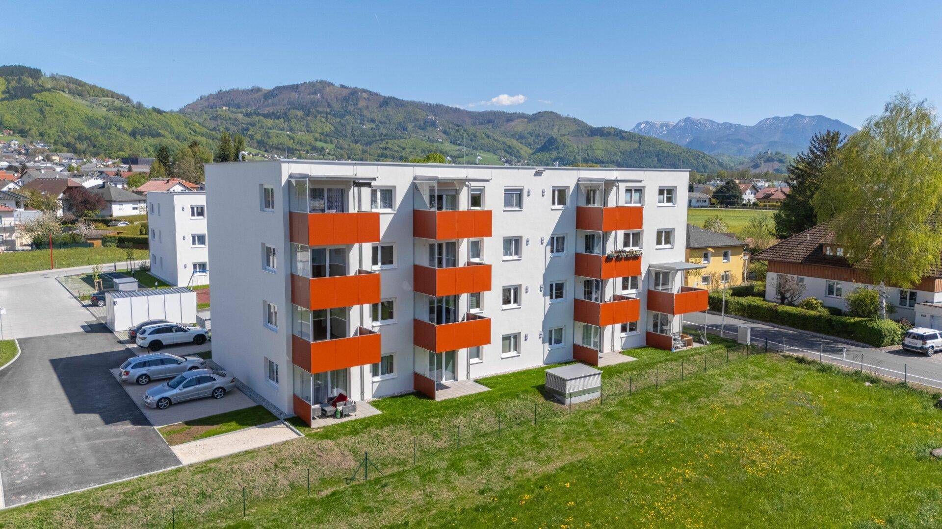 Neubau-Wohngebäude mit orangefarbenen Balkonen, umgeben von grünen Wiesen und Bergen unter blauem Himmel.