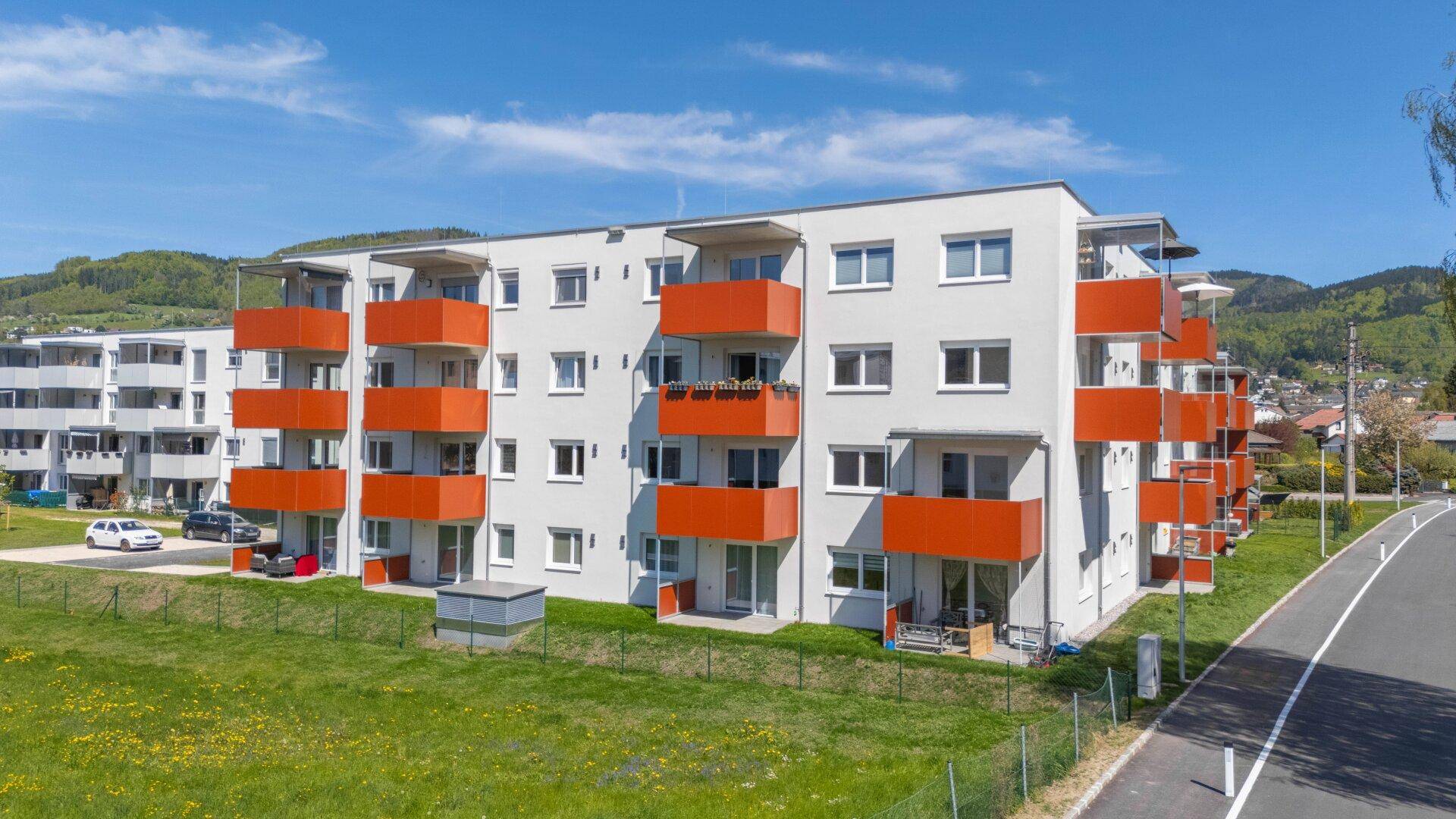 Modernes Mehrfamilienhaus mit leuchtend orangefarbenen Balkonen und Blick auf die umliegende Hügellandschaft.