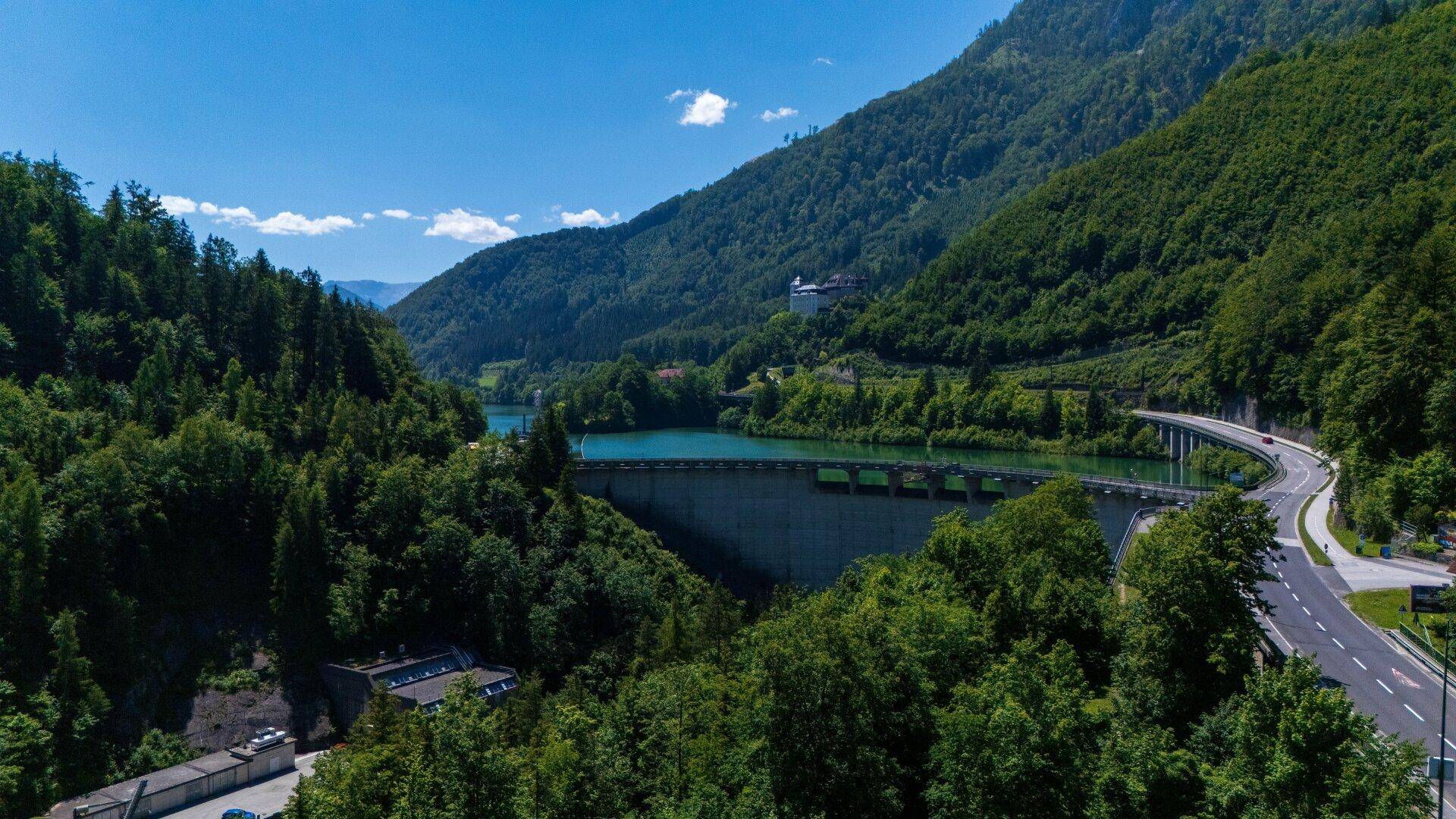 Panoramablick auf einen Staudamm und See in einer grünen Berglandschaft mit kurviger Straße.