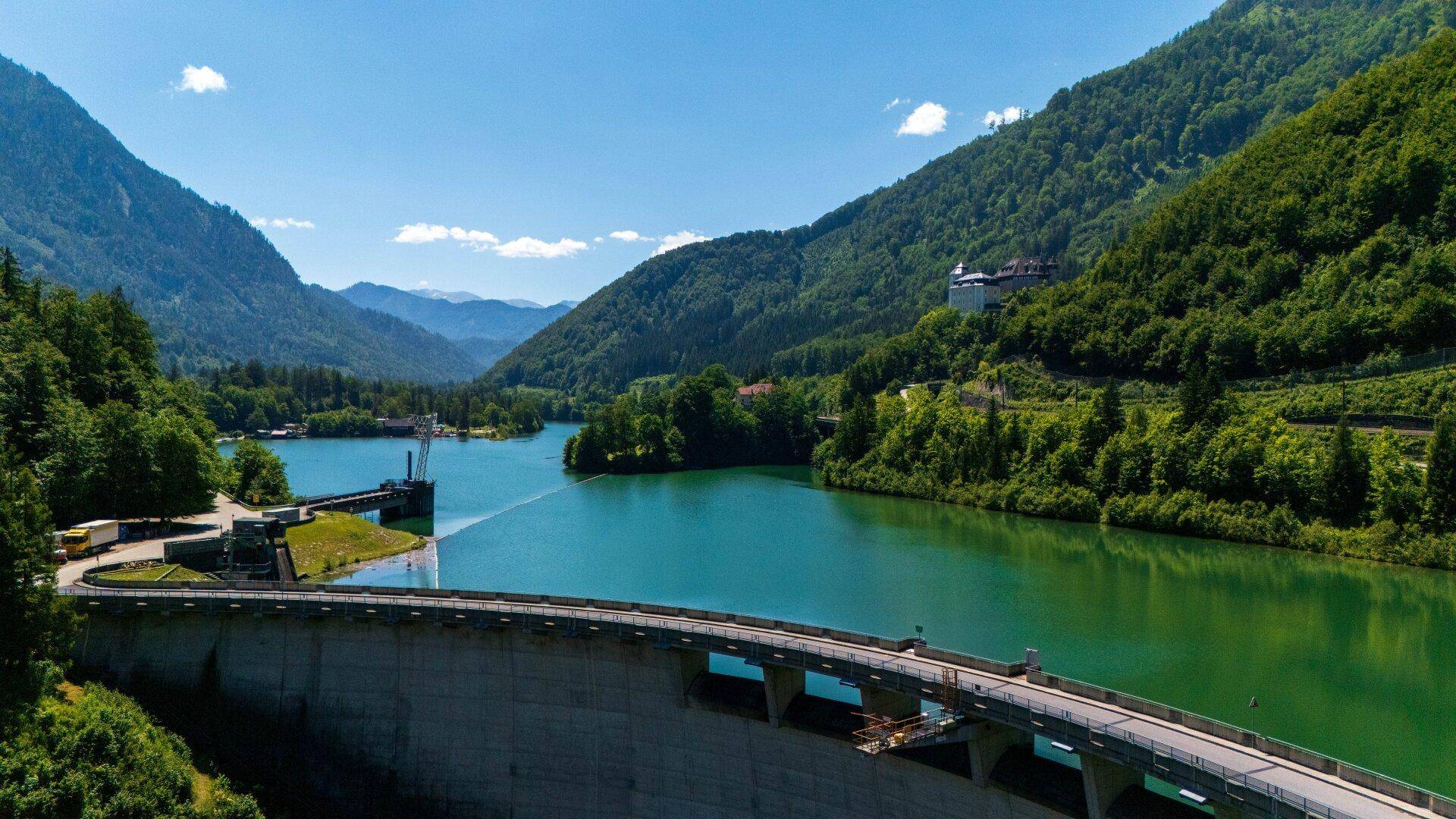 Panoramablick auf einen Staudamm mit türkisfarbenem See und bewaldeten Bergen im Hintergrund.