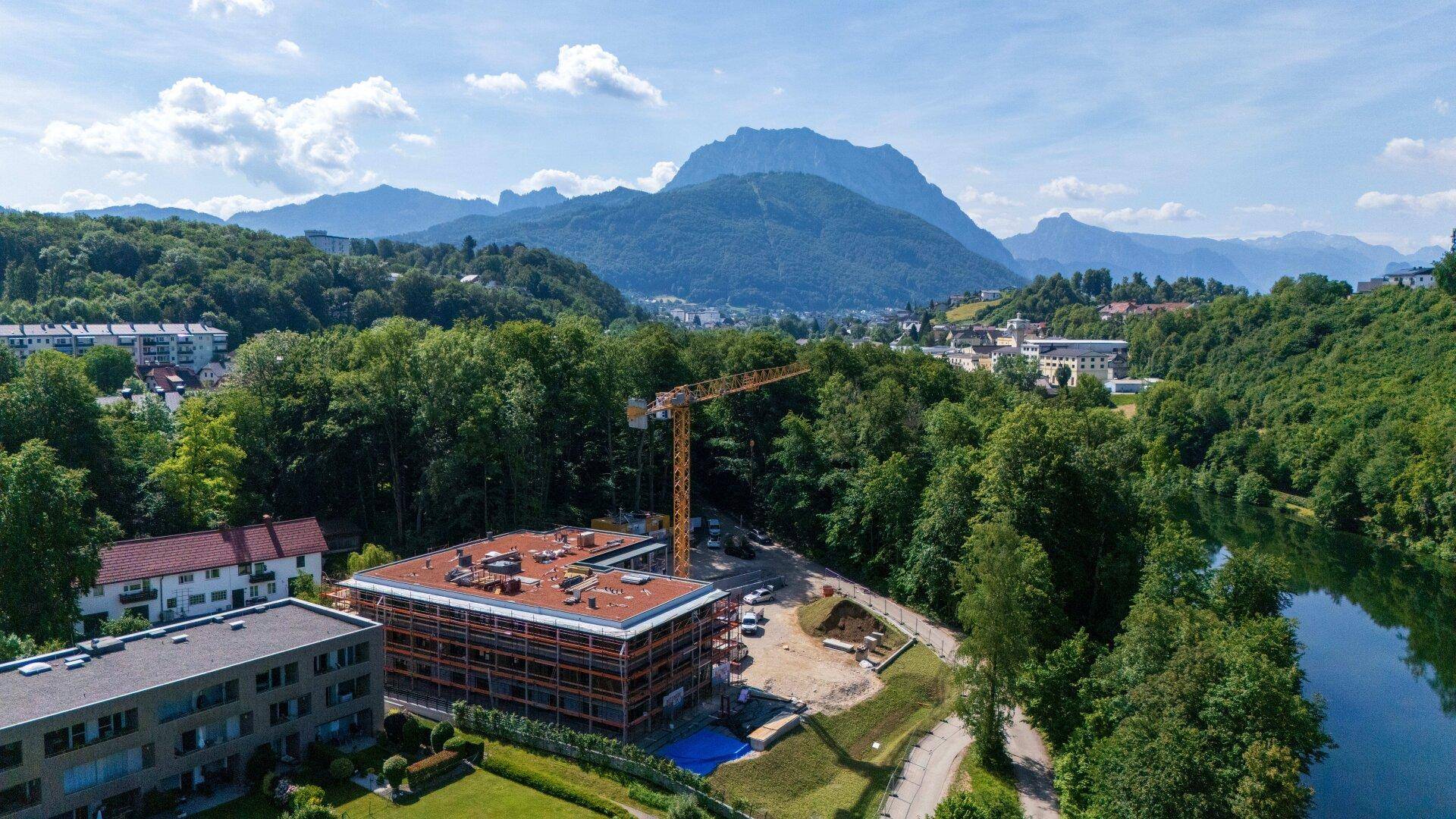 Luftaufnahme einer Baustelle am Flussufer mit umliegender Natur und Bergen im Hintergrund.