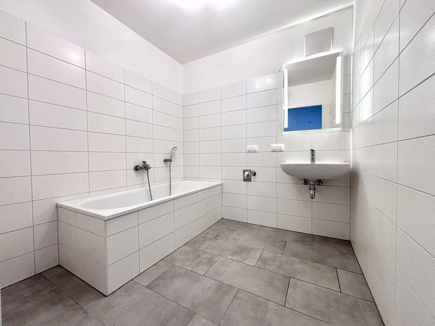 Modernes Badezimmer mit weißen Wandfliesen, grauen Bodenfliesen, Badewanne und beleuchtetem Spiegel.
