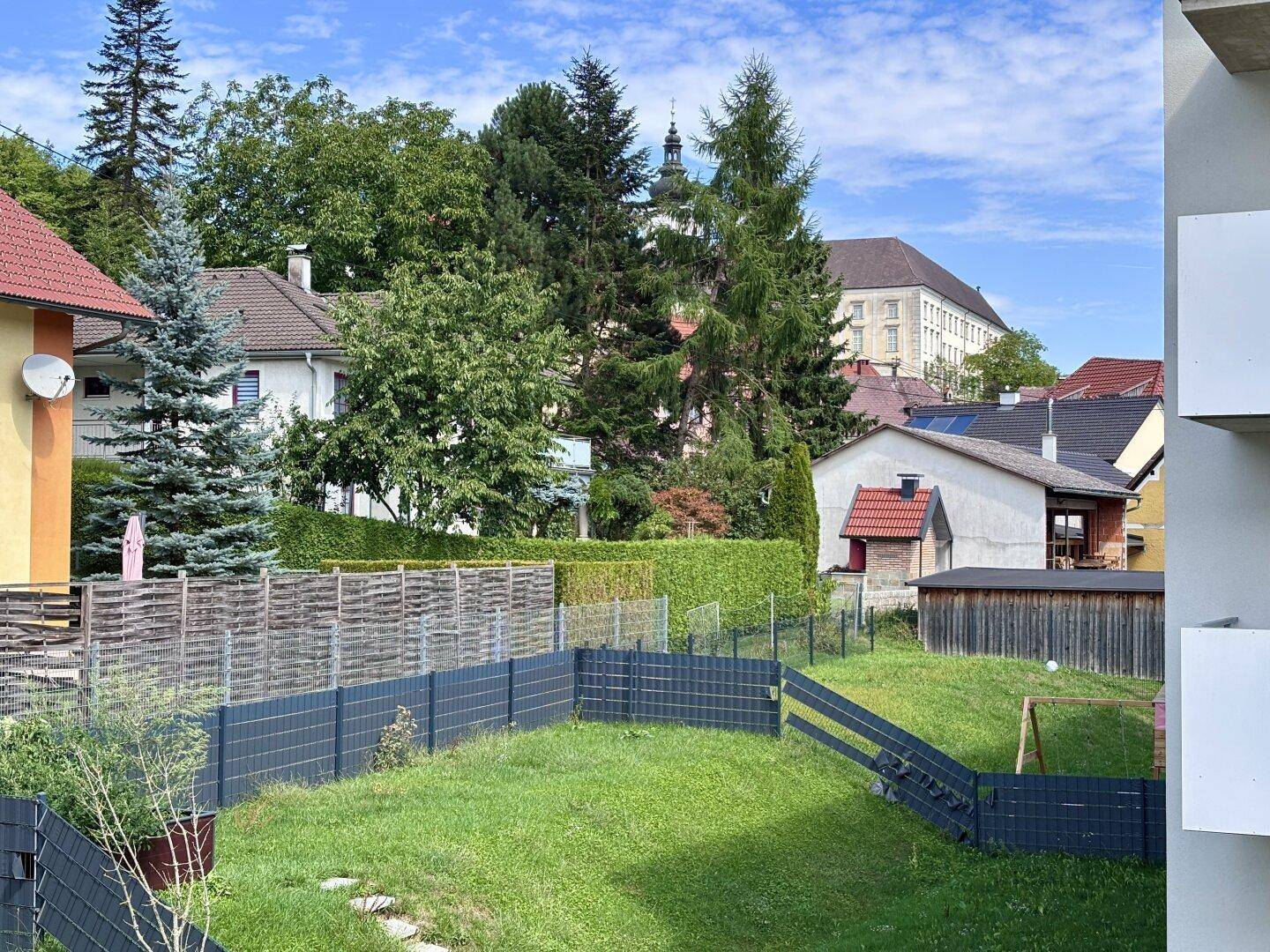 Blick auf einen gepflegten Gartenbereich mit Rasen, Zäunen und Bäumen im Hintergrund.