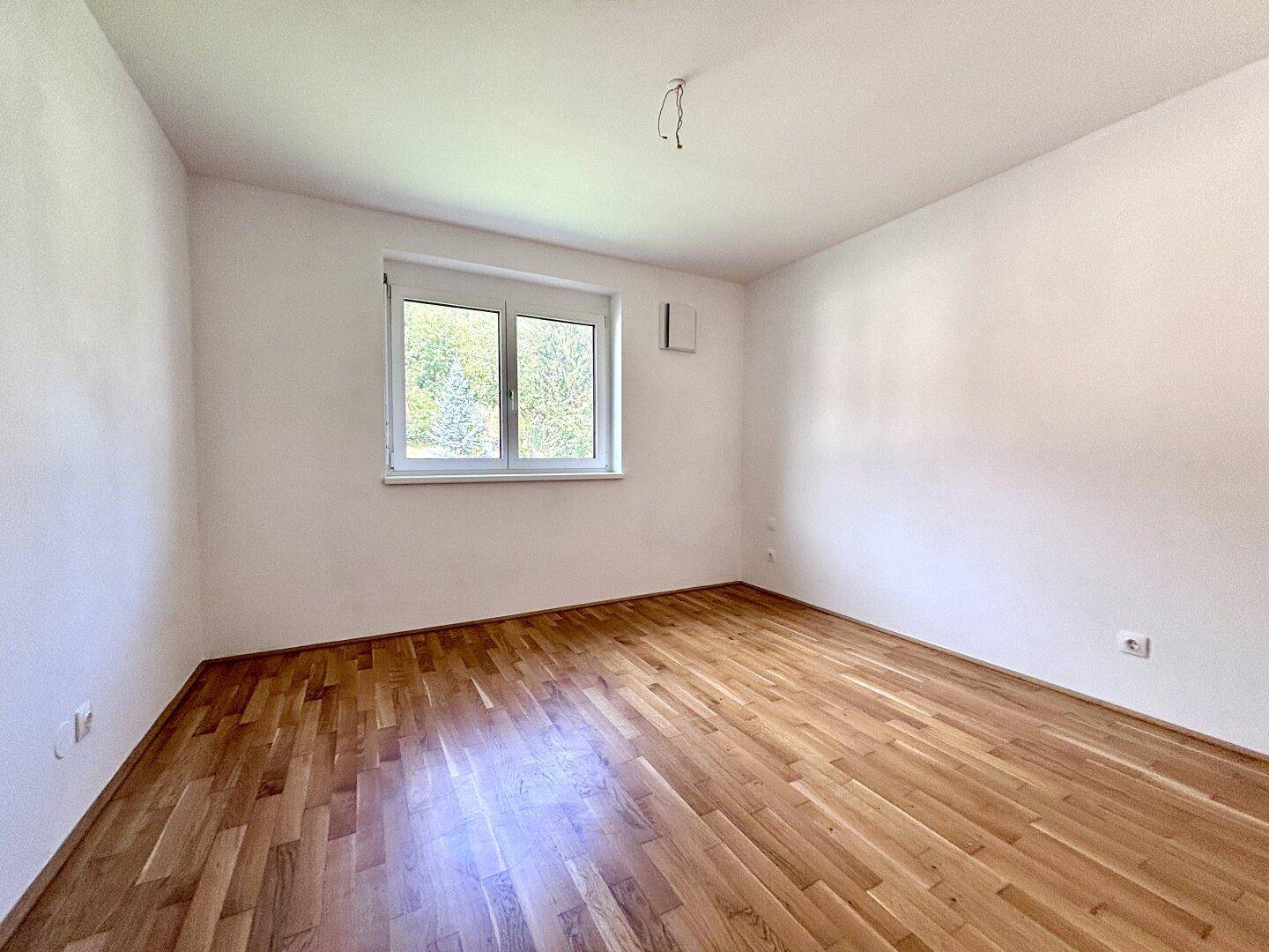 Ruhiges Schlafzimmer mit hellem Parkettboden und einem Fenster mit Blick ins Grüne.