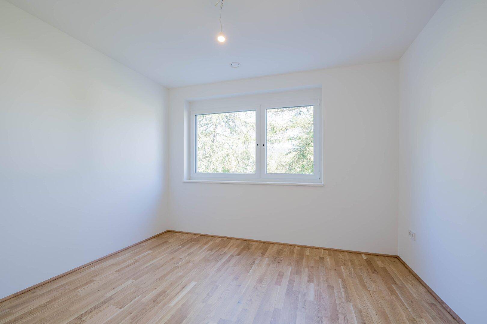 Geräumiges, helles Zimmer mit Holzfußboden und zwei Fenstern mit grüner Aussicht.