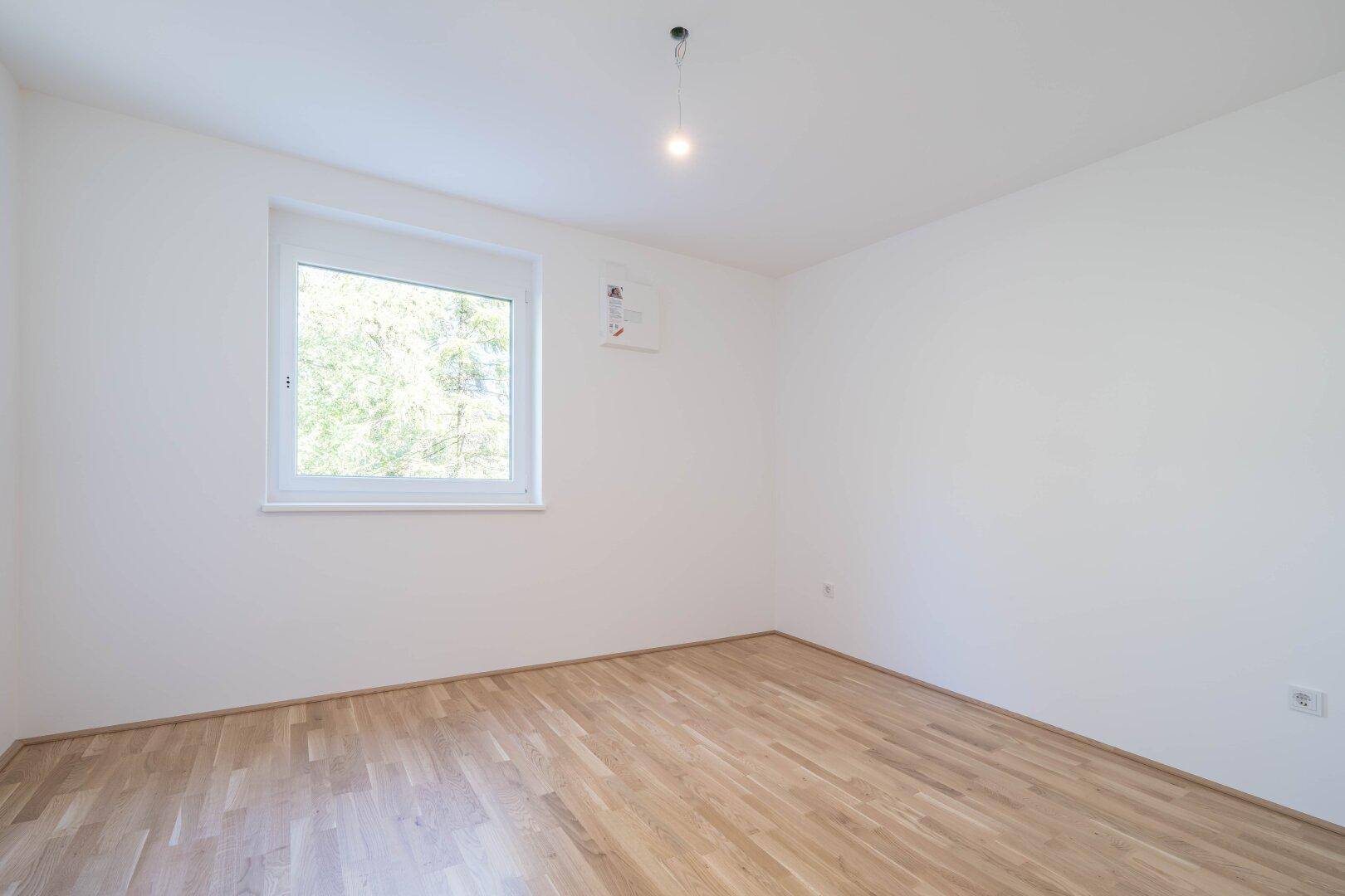 Helles Schlafzimmer mit Parkettboden und einem Fenster mit Blick ins Grüne.