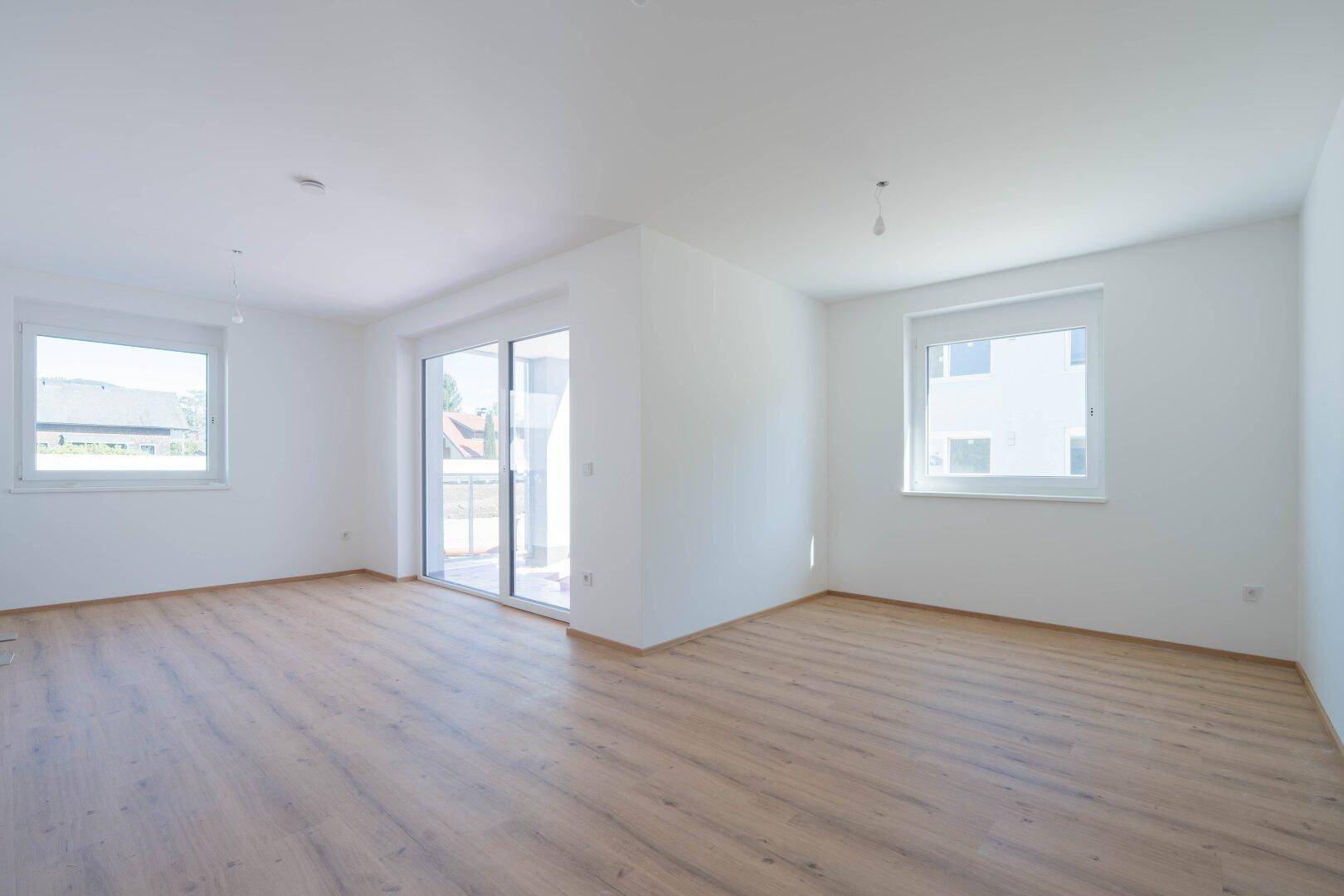 Geräumiger, heller Wohnbereich mit Laminatboden, großen Fenstern und Schiebetür zum Balkon.