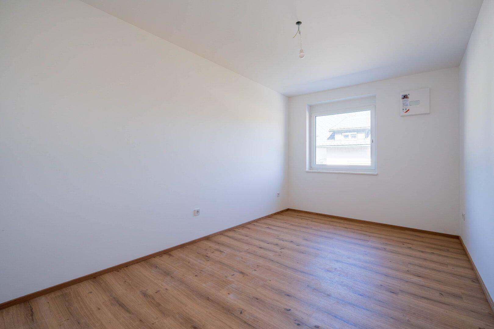 Leeres Schlafzimmer mit Laminatboden, weißen Wänden und einem Fenster, bereit zum Einzug.