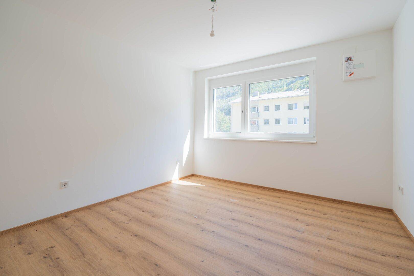 Helles Zimmer mit Laminatboden und Fenster, das einen Blick auf die umliegenden Gebäude und Natur bietet.