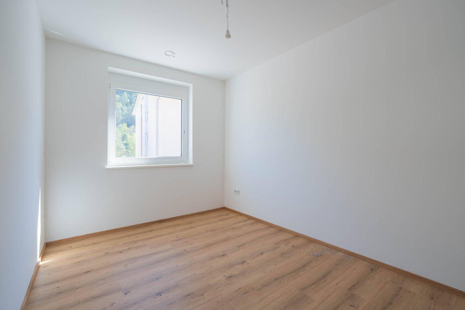 Schlafzimmer mit hellem Laminatboden, weißen Wänden und einem Fenster mit Blick ins Grüne.