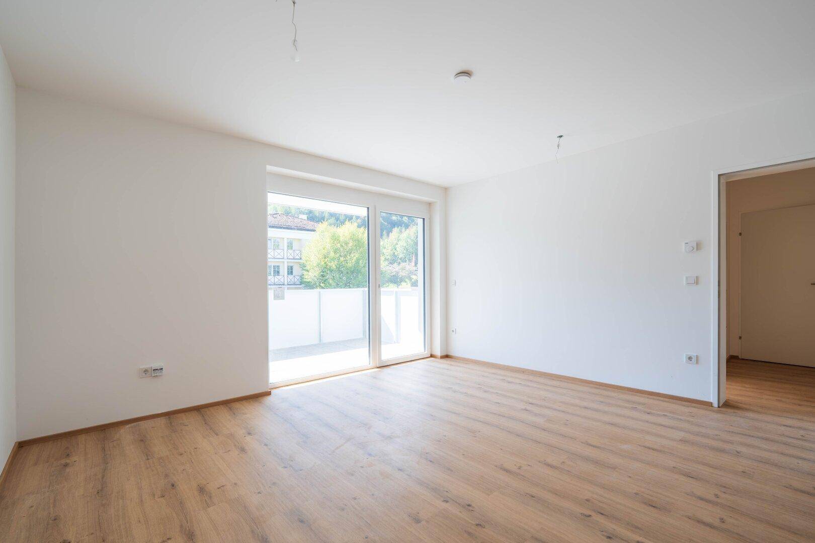 Heller, leerer Wohnraum mit Laminatboden und großem Schiebefenster zum Balkon.