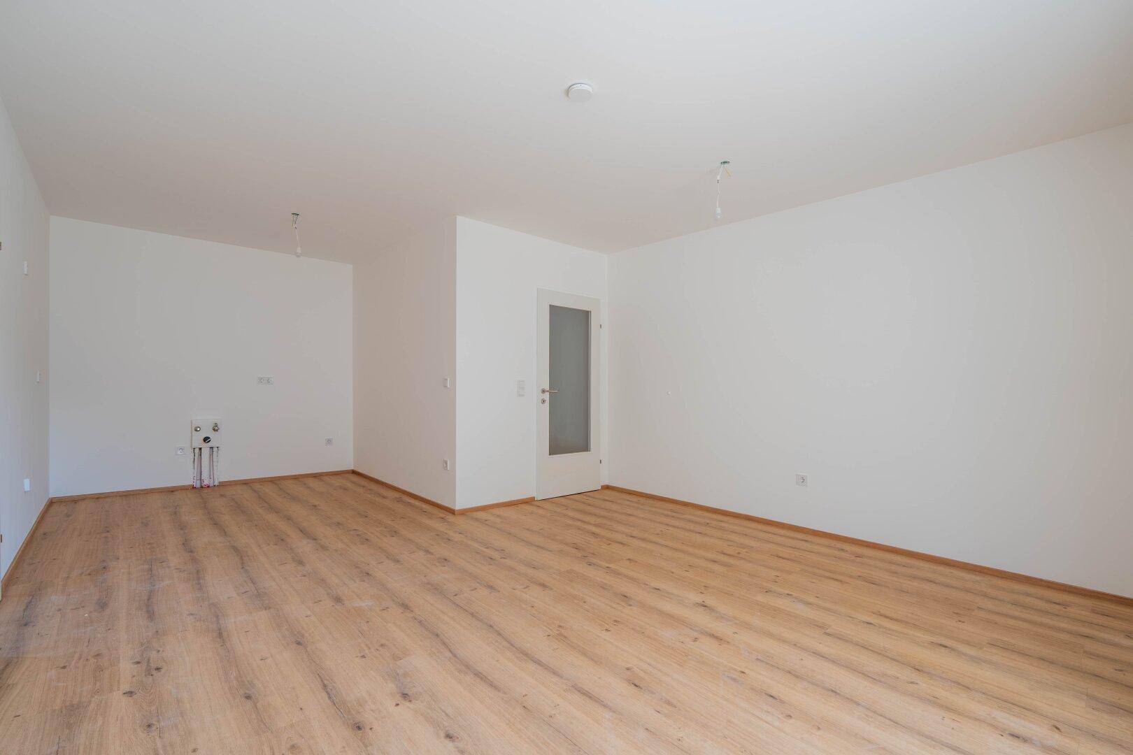Leerer Küchenbereich mit Laminatboden, weißen Wänden und Anschlüssen für eine Einbauküche.