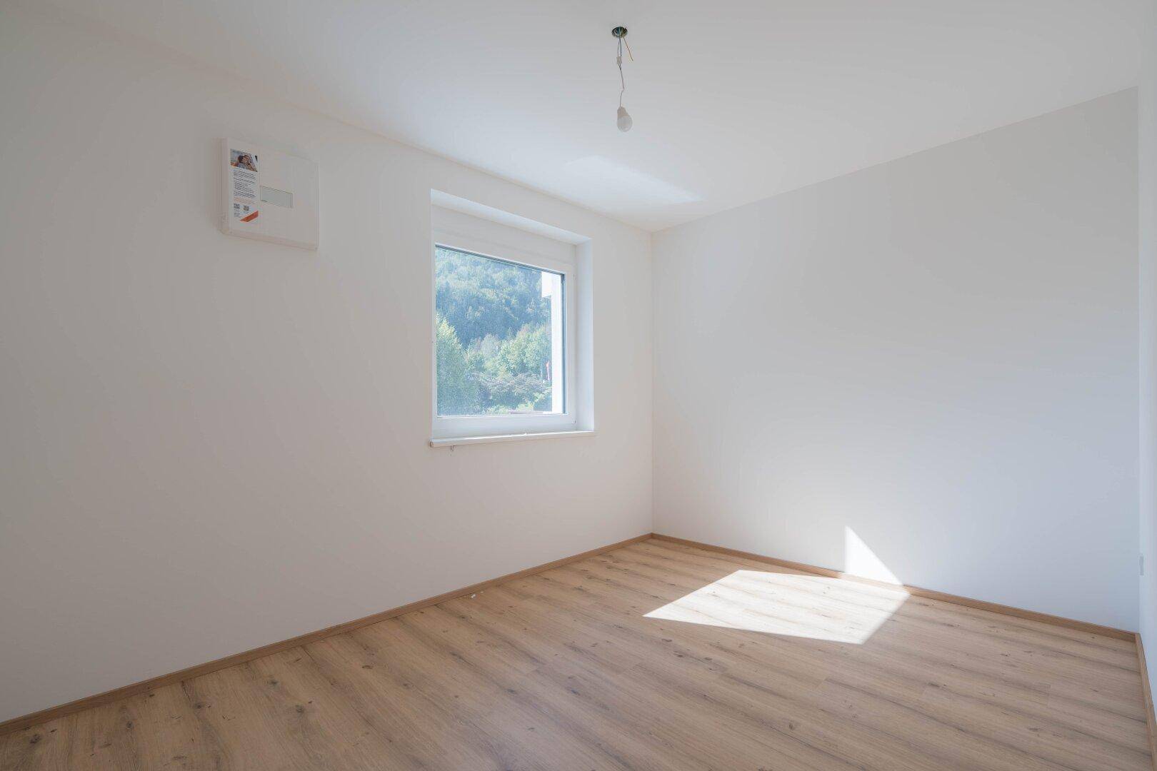 Helles Schlafzimmer mit Laminatboden, weißen Wänden und einem Fenster mit Blick ins Grüne.