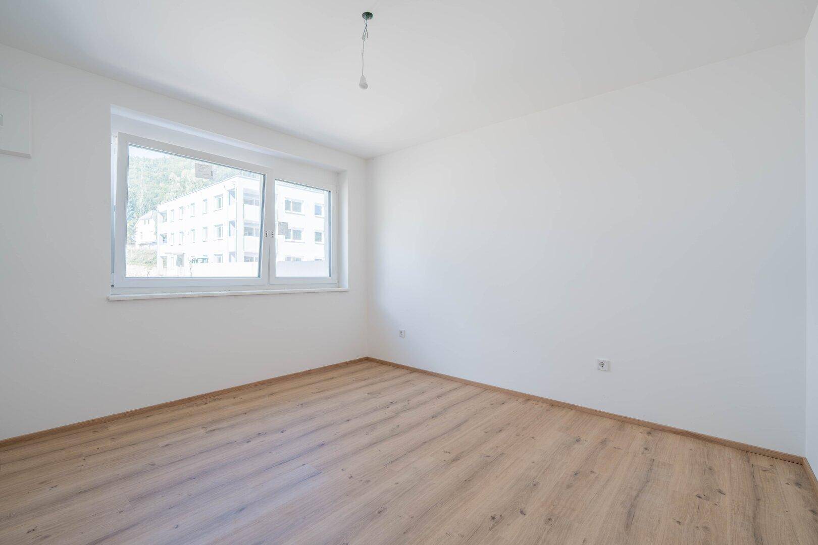 Leeres Schlafzimmer mit hellem Laminatboden und einem Fenster, das Tageslicht hereinlässt.