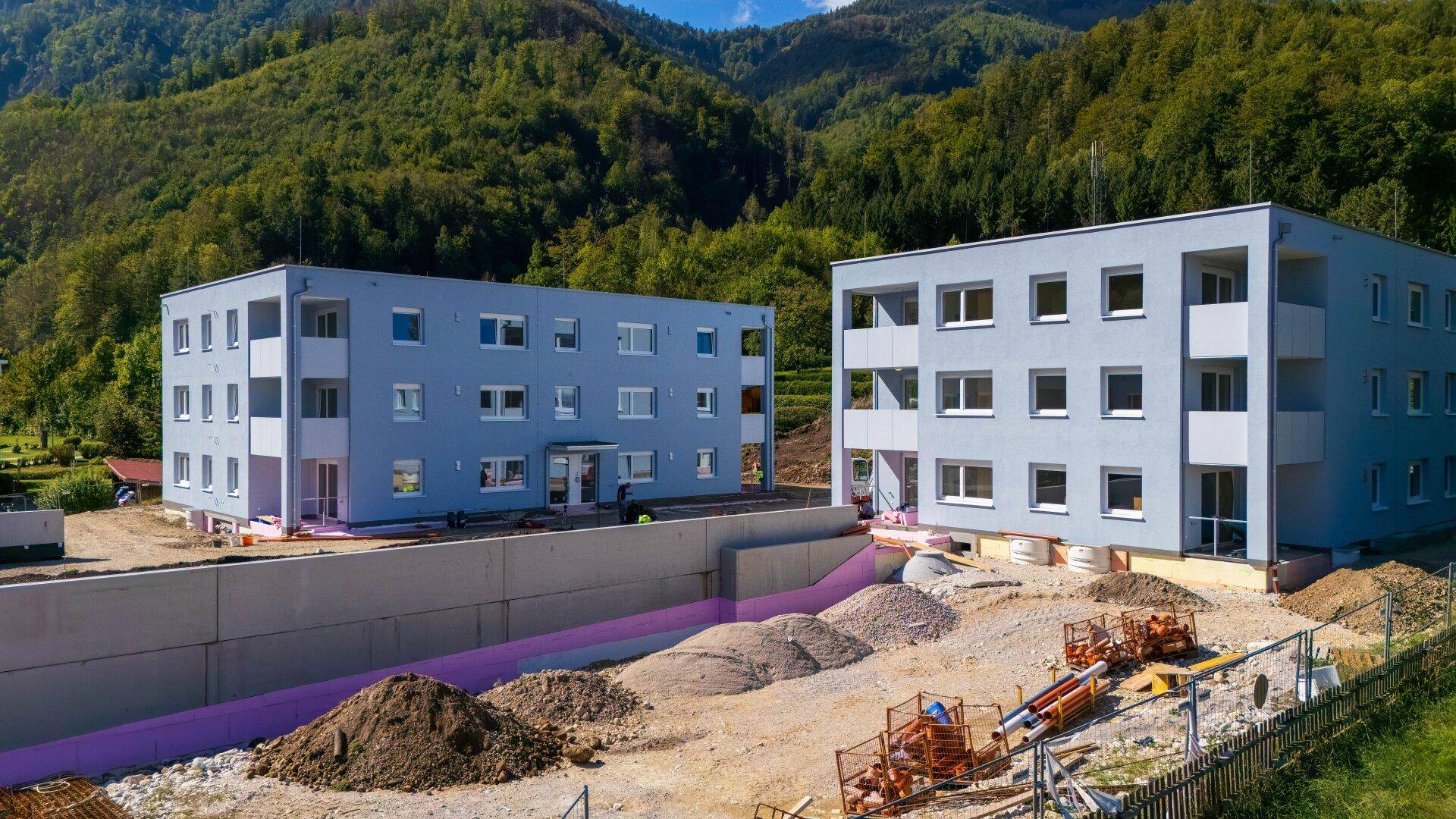 Zwei moderne Neubau-Mehrfamilienhäuser in einer grünen, bergigen Landschaft.