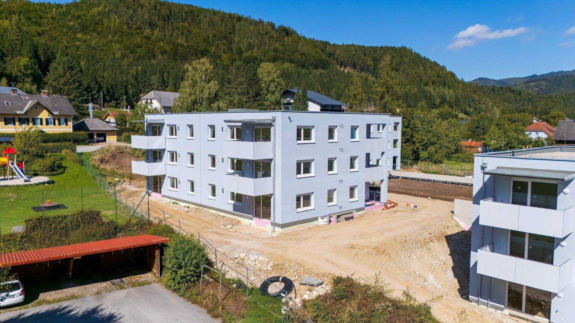 Modernes Mehrfamilienhaus in Neubauweise, umgeben von grüner Landschaft und anderen Wohnhäusern.
