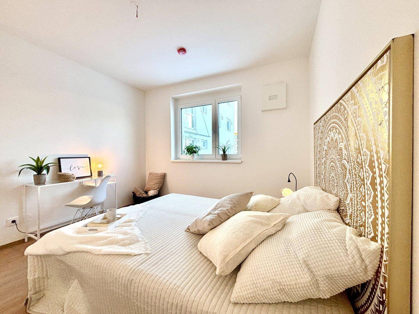 Gemütliches Schlafzimmer mit großem Bett, dekorativem Kopfteil und hellem Arbeitsbereich am Fenster.