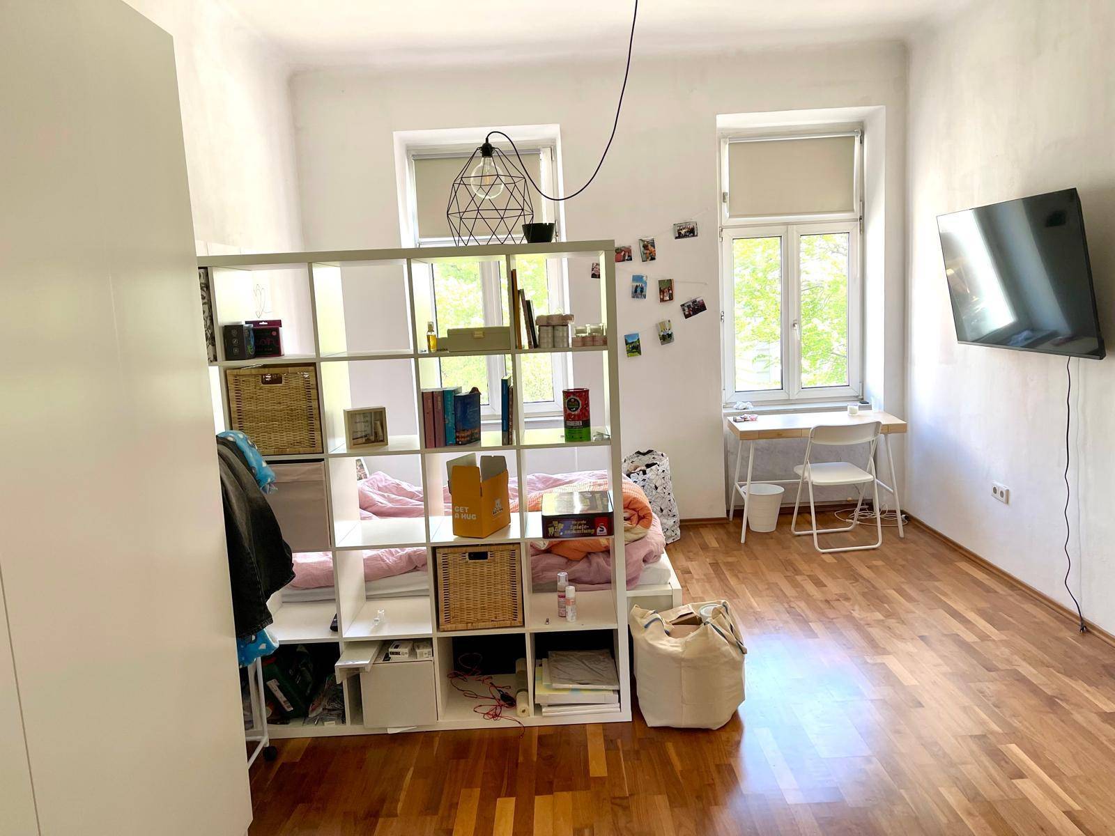 Helles Schlafzimmer mit Holzboden, Raumteiler und Schreibtisch am Fenster.