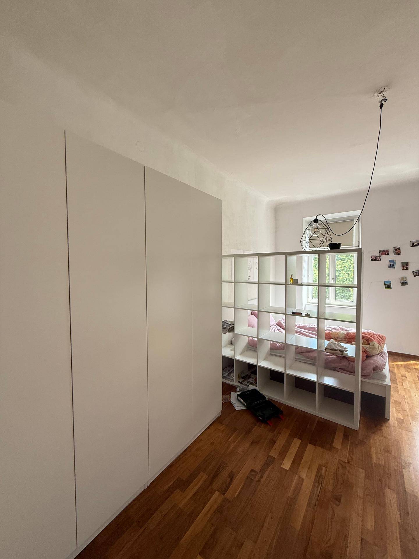 Schlafbereich mit weißem Raumteiler, Einbauschrank und Holzboden in einem hellen Raum.