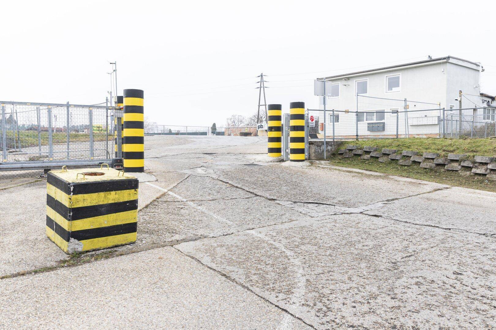 Zufahrt zu einem Gelände mit gelb-schwarzen Pollern und einem Gebäude im Hintergrund.